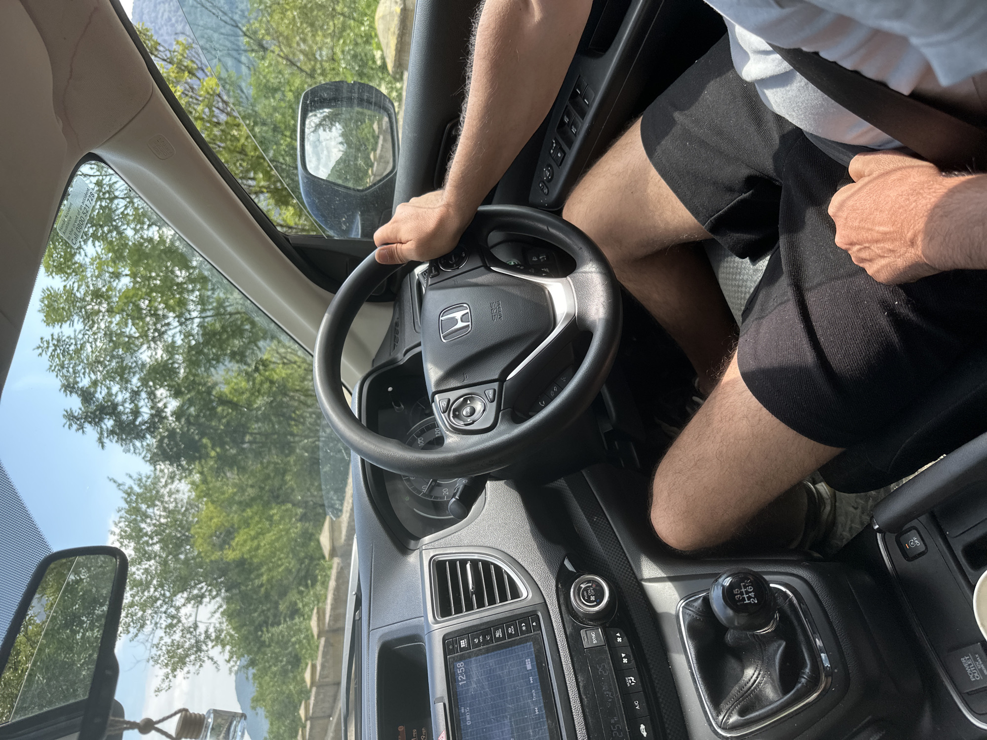 The image provides an interior view of a car from a high angle, focusing on the driver's area. An adult person, likely male, wearing a white t-shirt and black shorts, is seated in the driver's seat, secured by a seat belt. Their right hand is on the steering wheel, featuring a Honda logo, while their left arm rests near the manual gear stick.
The car's interior is gray and black, showing various dashboard elements. A central infotainment screen displays "12:58", "SYNC", and "25%", with climate controls below, including "A/C". A 6-speed manual gear stick is prominent, with the pattern "1 3 5 / 2 4 6 R" clearly visible. Other details include air vents, a "POWER OUTLET 120W MAX" socket, and a cup holder. The instrument cluster indicates "1 mpg" and "9 mph". An "SRS" airbag label is present on the A-pillar.
Through the windshield, a bright, sunny outdoor scene is visible, dominated by lush green trees and a clear blue sky. A stone or concrete barrier runs alongside the road, and the background hints at mountainous or hilly terrain. An air freshener hangs from the rearview mirror. A sticker on the windshield provides "Honda Accident Assistance" details, stating "In the event of an accident Call 0800 521 728". The bright lighting suggests it is daytime with good weather. The setting appears to be a scenic drive, consistent with the natural landscapes found around Drobeta-Turnu Severin, Romania.