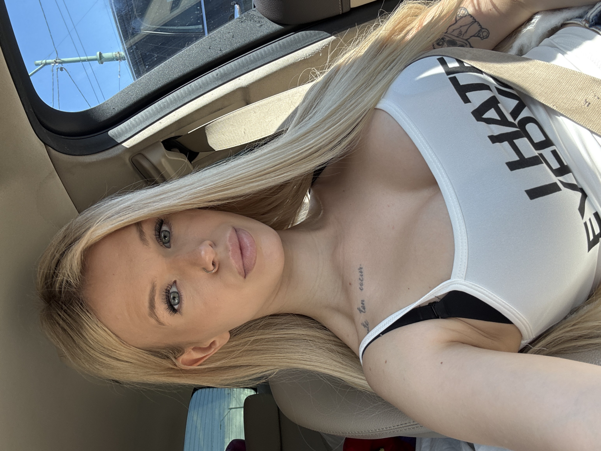 A blonde woman with blue eyes, a nose ring, and light skin is sitting in the driver's seat of a car. She is wearing a white tank top with black text that reads "I HATE EVERYBODY". There is a tattoo on her upper chest and another on her arm. The car appears to be parked on a street in Saint Petersburg, Russia, with a clear blue sky and a utility pole visible through the window. The sun is shining, casting shadows on the interior of the car.