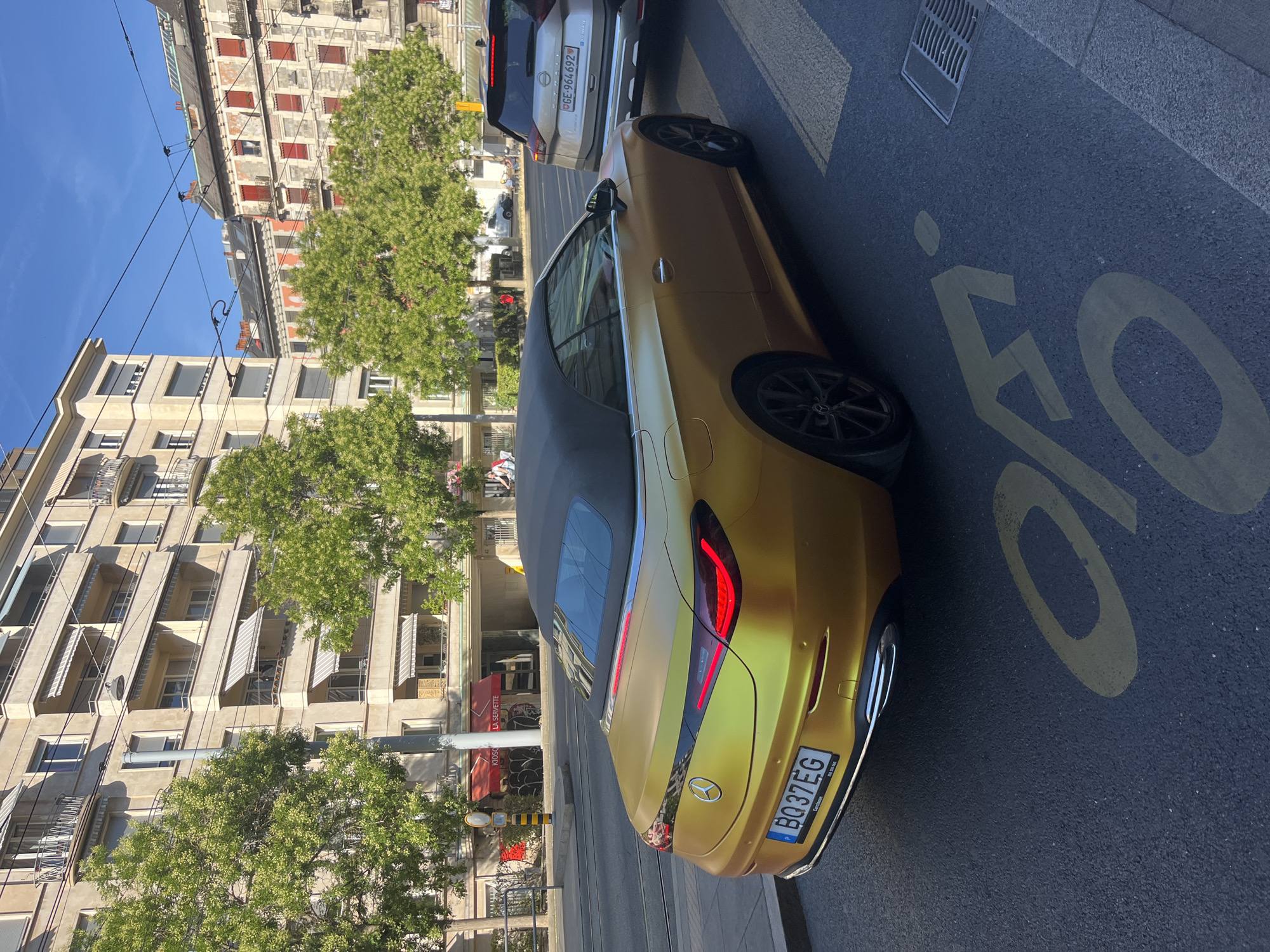 In Geneva, Switzerland, a gold Mercedes-Benz EQC convertible is parked on a street. The car has a black soft top and a license plate that reads "BQ37EG". It is parked next to a silver Nissan SUV with the license plate "GE-964692". The street has a bicycle lane marked with yellow paint. In the background, there are several multi-story buildings with awnings and trees lining the street. The sky is clear and blue, indicating it is a sunny day. A person is visible walking in the distance.