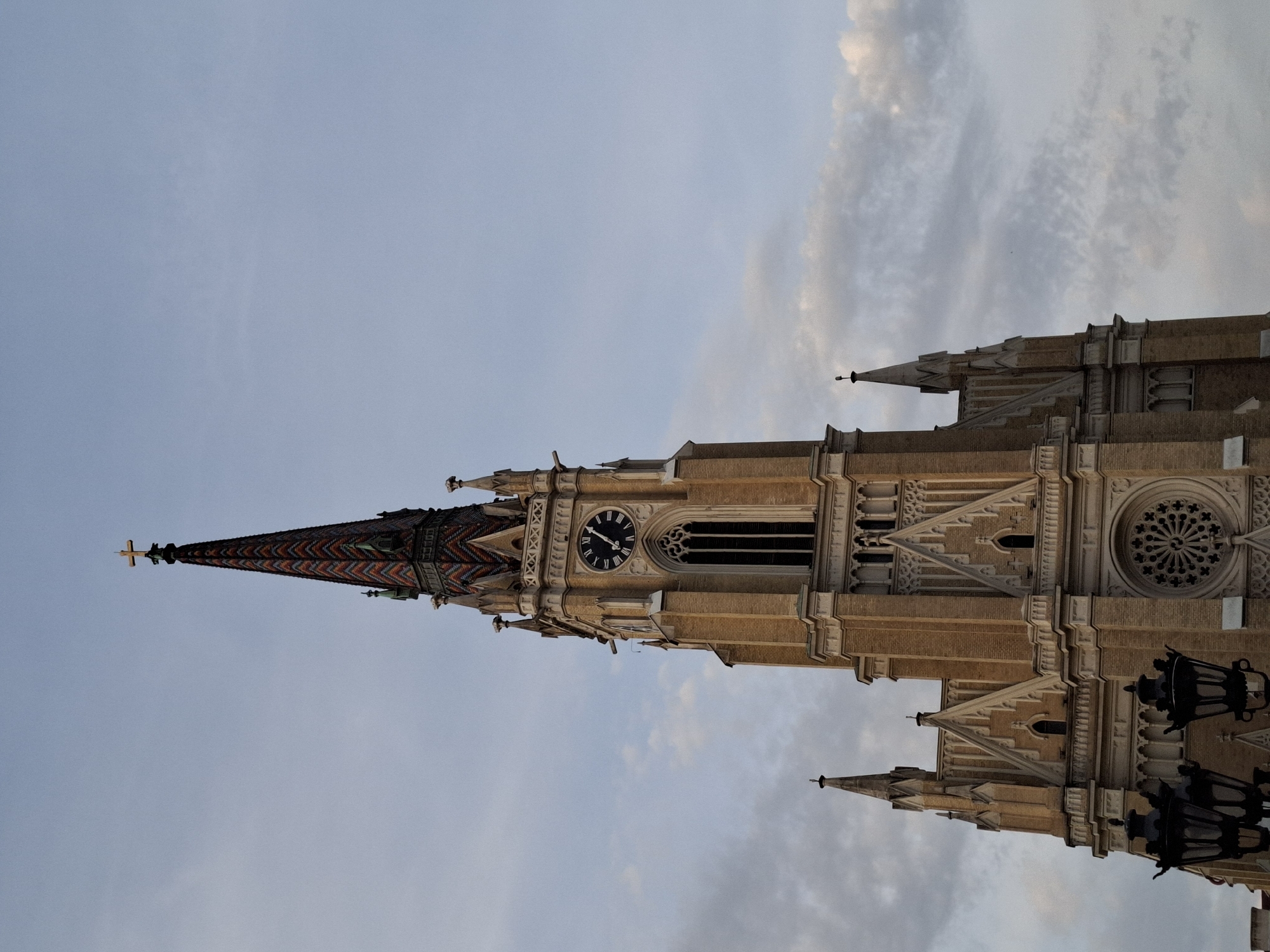 This striking image captures a vertical perspective of the Name of Mary Church in Novi Sad, Serbia, an impressive example of Gothic-revival architecture. The photograph is taken from a low angle, emphasizing the towering height and intricate details of the church's facade and spire against a soft, partly cloudy sky.
The central subject is the church's main tower, crowned with a remarkably tall, slender spire. The spire's roof features a distinctive, colorful tiled pattern, appearing as a herringbone or chevron design in shades of red, green, and dark blue or black. A golden cross adorns the very apex of the spire. Below the spire, a large, dark clock face with Roman numerals is visible, its hands indicating approximately ten minutes to six (5:50). The tower itself is constructed from light brown or beige brick or stone, contrasted with lighter, ornate decorative elements such as cornices, buttresses, and intricate window tracery. A prominent rose window, a characteristic feature of Gothic architecture, is visible on the upper facade to the right of the clock tower. Smaller spires and architectural details extend from the main structure, adding to its grand and complex silhouette.
The sky is a soft blue-grey, suggesting either late afternoon or early evening, with scattered, wispy clouds that create a diffused, ambient light. There are no people visible, and the scene is static, focusing entirely on the architectural splendor of the building. In the bottom right corner, a part of an ornate black streetlight or lantern is partially visible. The only visible text consists of the Roman numerals on the clock face: I, II, III, IV, V, VI, VII, VIII, IX, X, XI, XII.