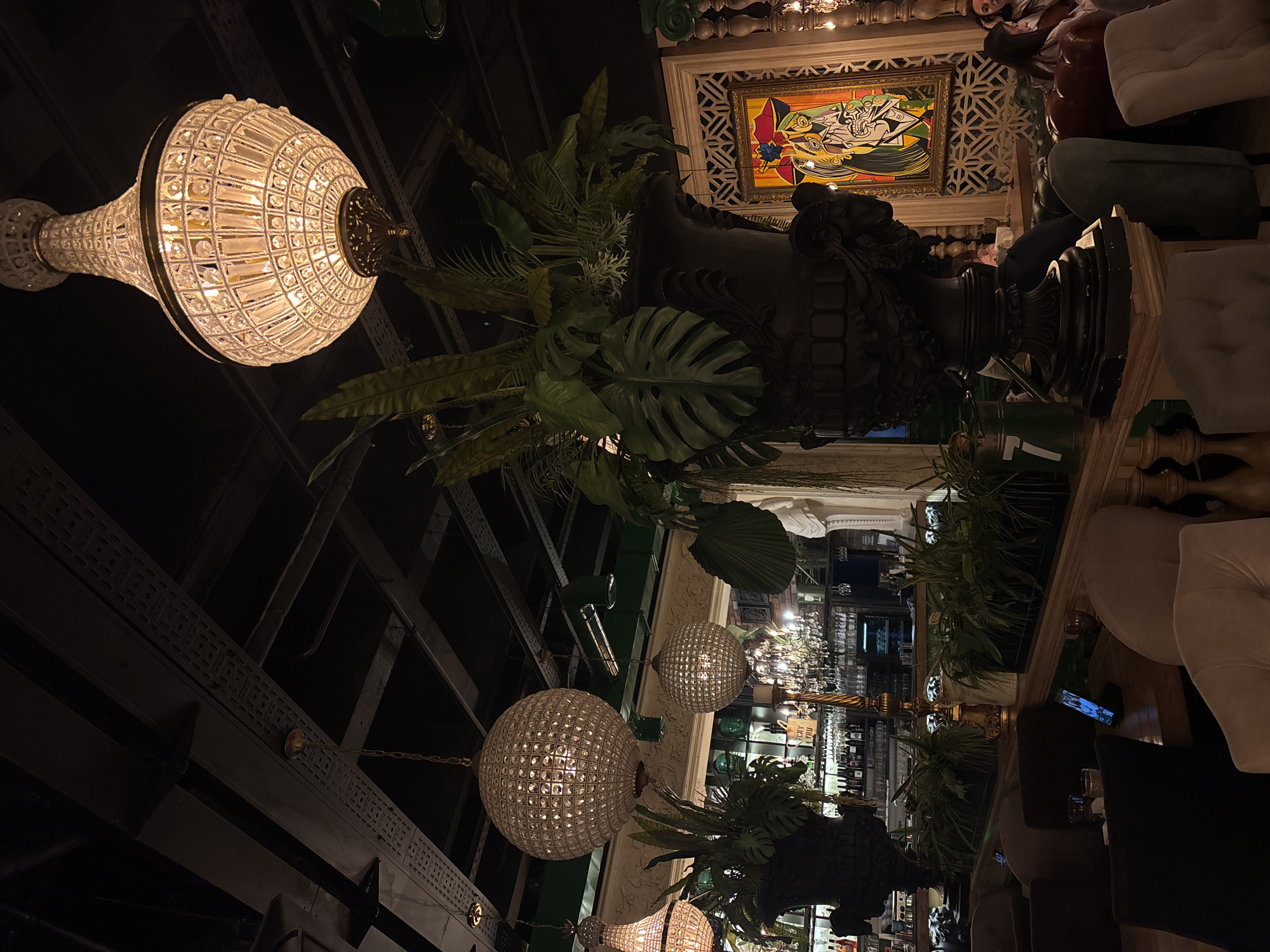 The image captures the interior of a bar or restaurant in Saint Petersburg, Russia. The scene is dimly lit, showcasing an opulent atmosphere. Several crystal chandeliers hang from the ceiling, providing a warm glow. Large, dark-colored urns filled with lush green plants decorate the space, adding a touch of nature to the elegant setting. In the background, a bar area is visible, lined with shelves of bottles. Tables and chairs are arranged for patrons, and a framed artwork hangs on the wall. People are seated and enjoying the ambiance.