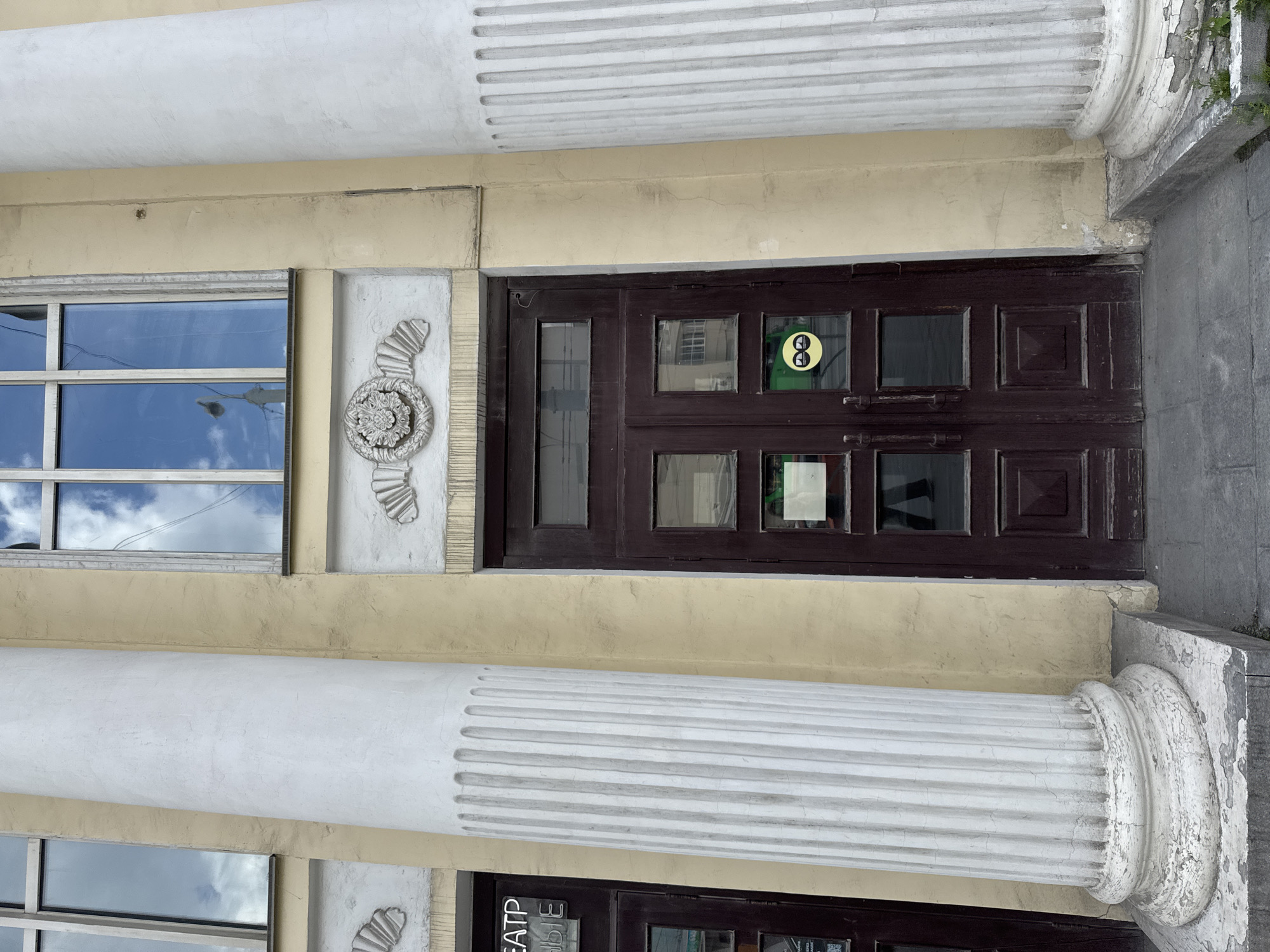 The image shows the entrance to a building in Yekaterinburg, Russia. The entrance features a set of dark brown double doors with glass panes. Above the doors is a decorative element with a circular floral motif and shell-like carvings on either side. Two large, fluted white columns flank the entrance. One column is on the left, partially obscuring a door with visible text that reads "ТЕАТР" and another line below it. The other column is on the right, positioned closer to the main double doors. Above the entrance, there are tall windows with reflections of the sky and clouds. The building's facade is painted a pale yellow. On the right-hand door, there is a round yellow sticker with a black graphic of two figures wearing glasses, facing each other. The ground in front of the entrance is paved with gray stone slabs. The overall scene depicts the exterior of a public building, likely a theater or cultural institution, in an urban setting.