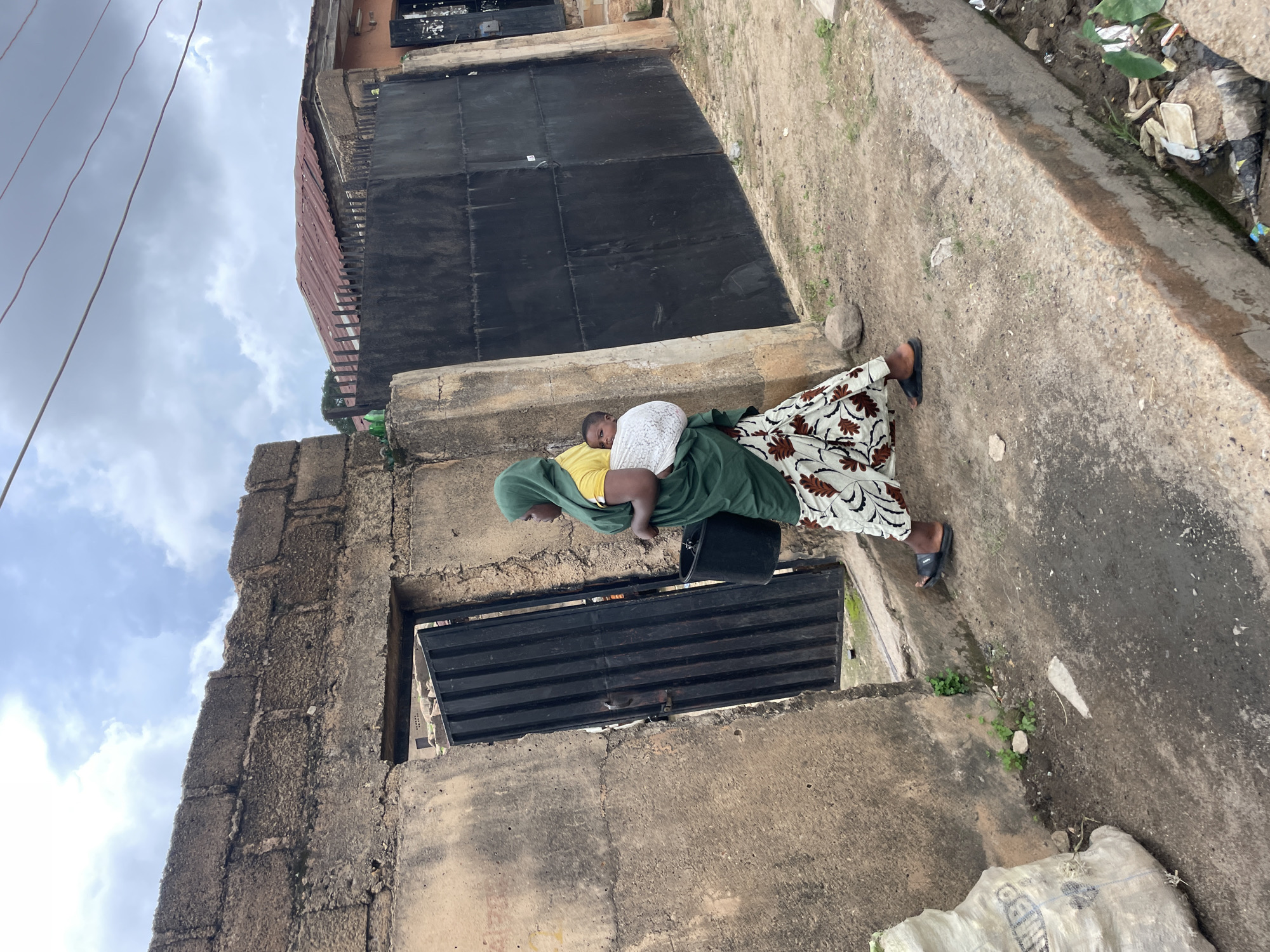 The image shows a woman carrying a baby on her back, walking on a dirt path next to a building. The woman is wearing a green headscarf and a patterned dress. The baby is wrapped in a white cloth. The building appears to be made of concrete blocks and has a corrugated metal roof. A large black metal gate is visible on the building. There is some trash on the ground near the building. The sky is cloudy. The scene appears to be in a residential area. The location is Jos, Nigeria. There is no visible text.