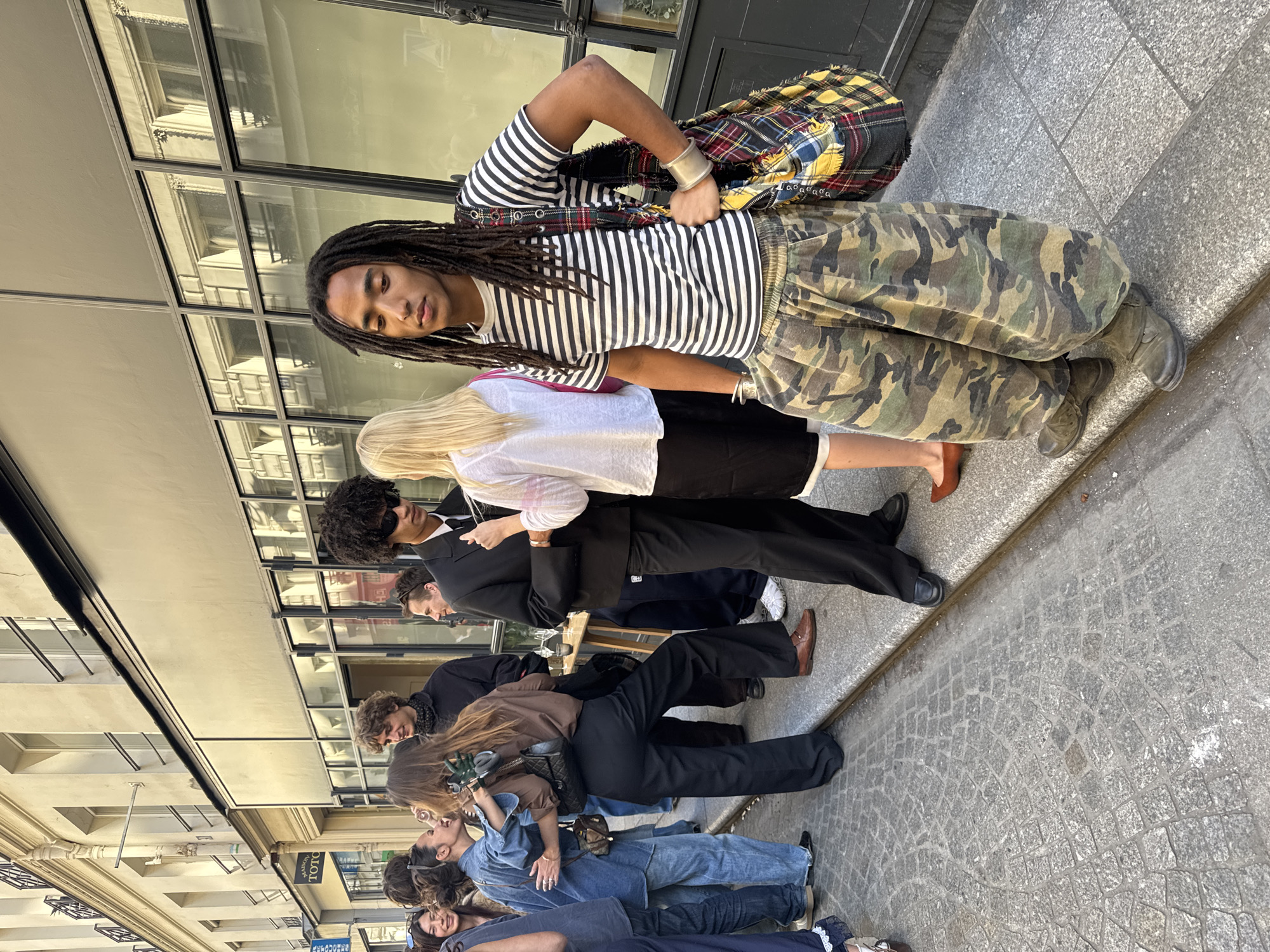 An outdoor street scene, rotated vertically, captures a diverse group of people in what appears to be a European city. The primary subject, a person with long dreadlocks, stands in the foreground, looking directly at the viewer with a neutral expression. They wear a black and white striped short-sleeve t-shirt, baggy camouflage cargo pants, and dark boots. A colorful plaid patchwork bag is slung over their shoulder, and they wear a large silver cuff bracelet on their left wrist.
Behind and to the side of this individual, several other people are visible. A blonde person in a white t-shirt is partially obscured. Further back, a group of people is gathered, some appearing to be conversing or walking. Their attire ranges from casual (denim, t-shirts) to semi-formal (black suits, dress shirts). Notable figures include a person in a black suit and sunglasses, and another in a denim jumpsuit.
The setting features multi-story buildings with large windows, reflecting the architecture across the street and the bright daytime sky. The street itself is paved with grey cobblestones and larger flagstones. The sun is shining brightly, creating distinct shadows, suggesting a clear day. A small, white sign with the text "TOTO" in dark letters is visible on one of the buildings in the mid-ground. The overall impression is a bustling urban environment, likely in Paris, given the architectural cues and provided context.