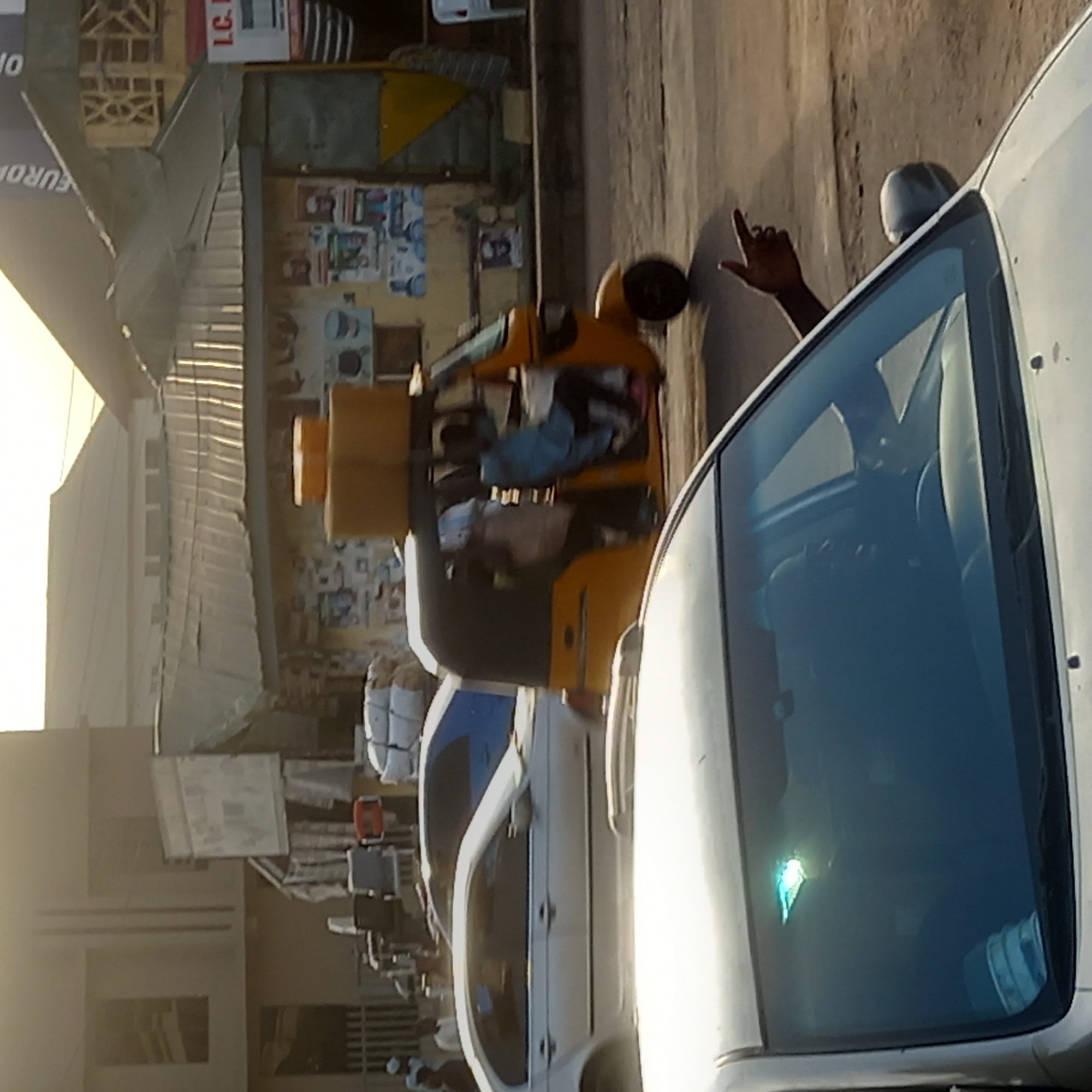 The image shows a busy street scene in Dadi Riba, Nigeria. A white car is in the foreground, with a man's hand visible through the windshield. Next to the car, there is a yellow tricycle loaded with goods, including stacks of yellow containers and what appears to be sacks of white cotton material. The street is lined with buildings and other vehicles. The overall impression is of a lively, bustling urban environment.