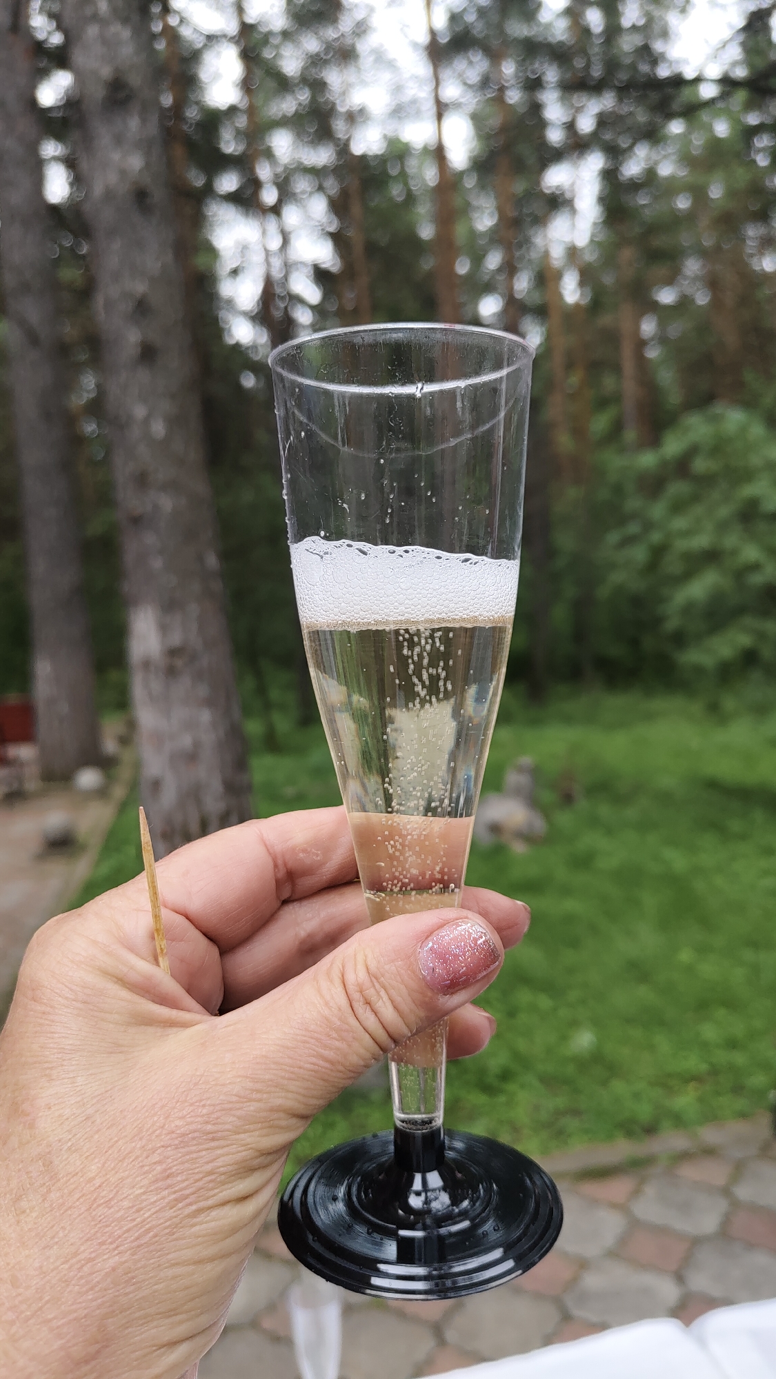 A person's hand with pink, glittery fingernails holds a champagne flute filled with a light yellow, bubbly beverage. The glass has a black base and is a plastic disposable type. A small wooden toothpick is also visible protruding from between the fingers. The background shows a blurred forest scene with tall trees and green foliage, suggesting an outdoor setting, possibly in or near Beryozovka, Russia. The ground is paved with hexagonal tiles.