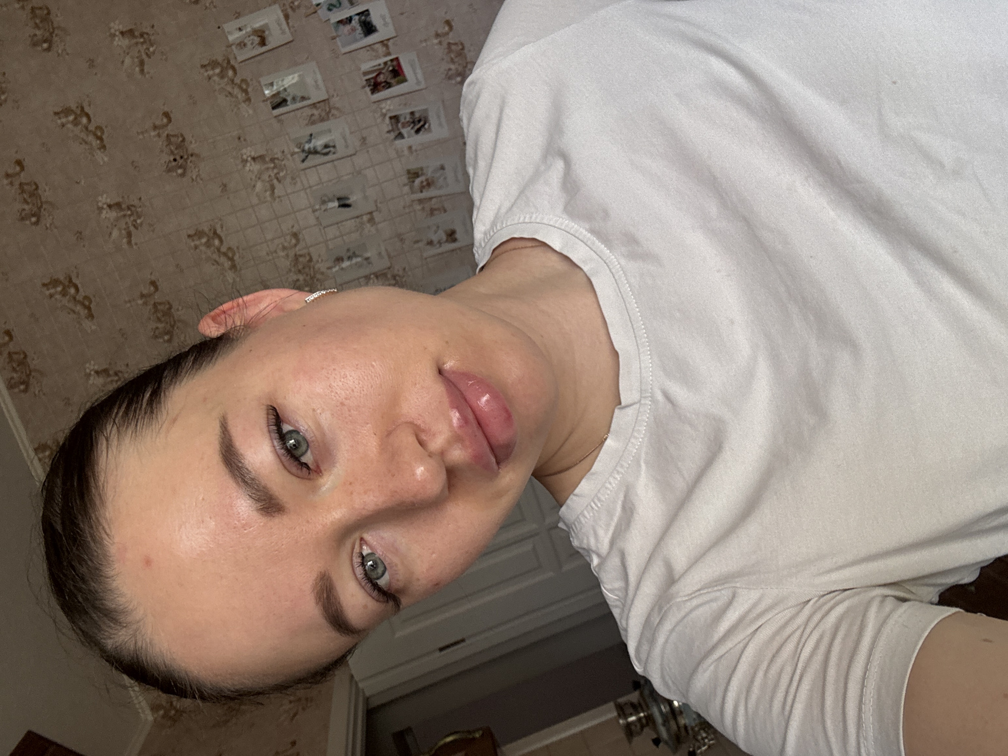 A woman with green eyes and dark hair is shown in a close-up, slightly tilted, indoor shot. She wears a white t-shirt. The background reveals a patterned wallpaper with small picture frames and glimpses of a doorway and light fixtures. The location is identified as Ферма, Russia.