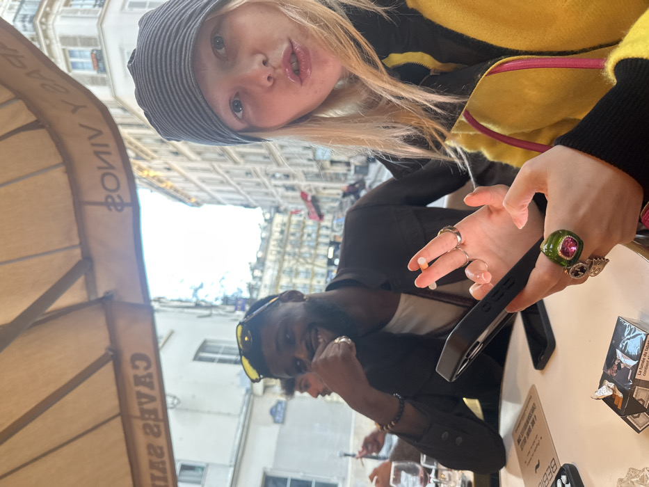 The scene depicts three individuals at an outdoor cafe terrace in Paris, France. In the immediate foreground, to the right, a person with blonde hair, wearing a grey striped beanie and a yellow and black jacket, holds a black smartphone in their left hand and an unlit cigarette between the fingers of their right hand. Multiple rings are visible on their hands, including a prominent green and pink ring on the left ring finger. Their lips are slightly parted, forming a pursed expression, and they look towards the camera. To their left, a Black male with a beard, wearing yellow sunglasses on his head, a white shirt, and a dark brown jacket, smiles towards the camera with his left hand near his mouth, displaying rings on his fingers. A third male is partially visible further left, holding a cigarette. The white table surface holds a menu, a pack of cigarettes (featuring French text), an ashtray containing crumpled foil, and at least two glasses of red wine. Above the patrons, a beige awning displays mirrored French text, including "CAVES SALLE" and "PARIS Y VINS." The background reveals a classic Parisian street scene with multi-story Haussmannian buildings and a blue street sign under an overcast sky.