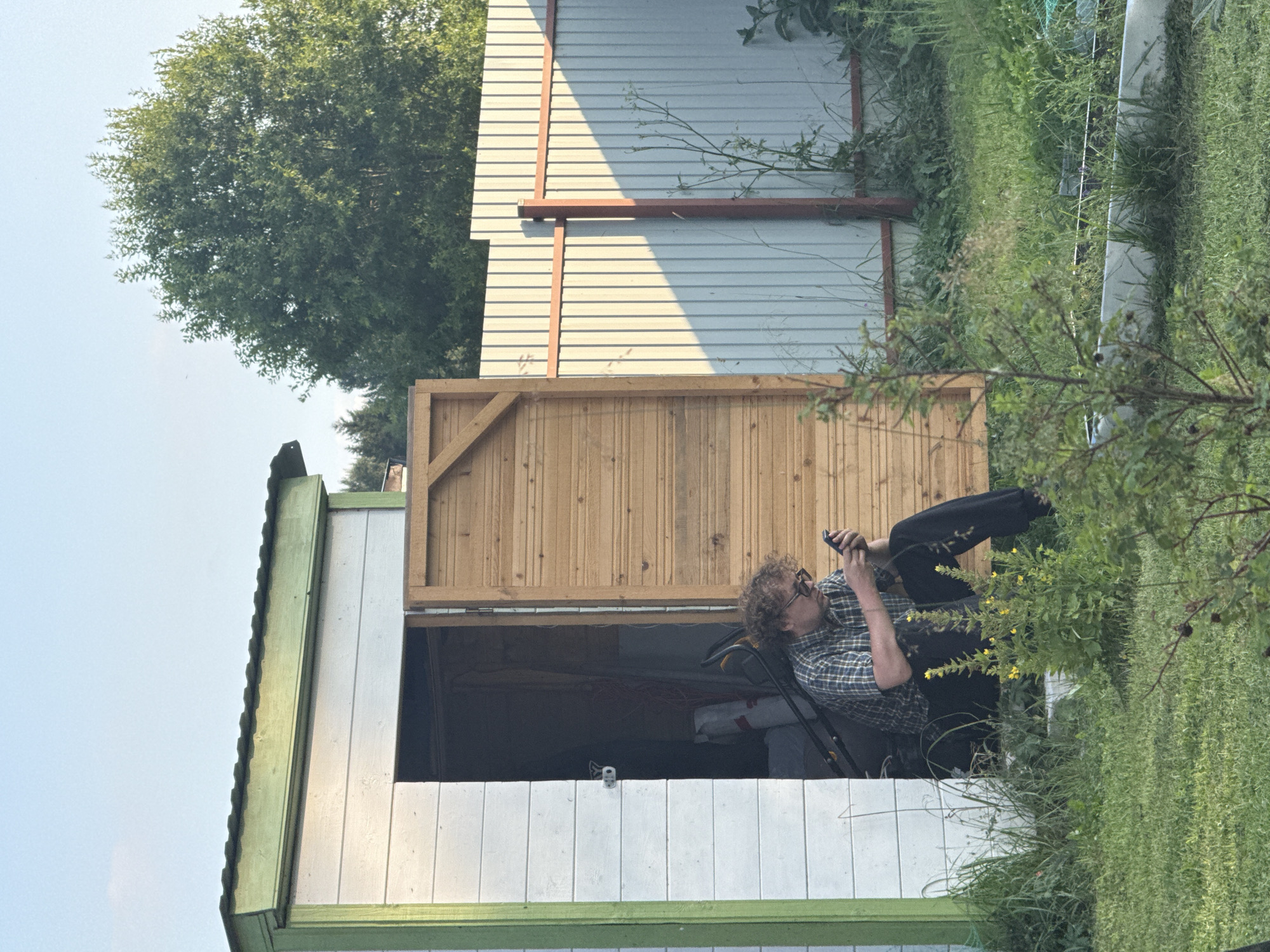 A young Caucasian man with curly blond hair and glasses is sitting in the open doorway of a white wooden shed with a green trim. He is wearing a plaid shirt and dark pants. He is looking at and holding a dark-colored smartphone in his hands, which he appears to be using. The shed's wooden doors are open. Inside the shed, a lawnmower and some bags are visible. The shed is situated in a grassy yard. A light-colored corrugated metal fence runs along the right side of the yard, with a wooden fence to the left of the shed. Trees are visible in the background. The scene appears to be in a rural or suburban area, potentially the city of Lyady, Russia, given the context. The lighting suggests it is daytime, possibly late afternoon due to the shadows.