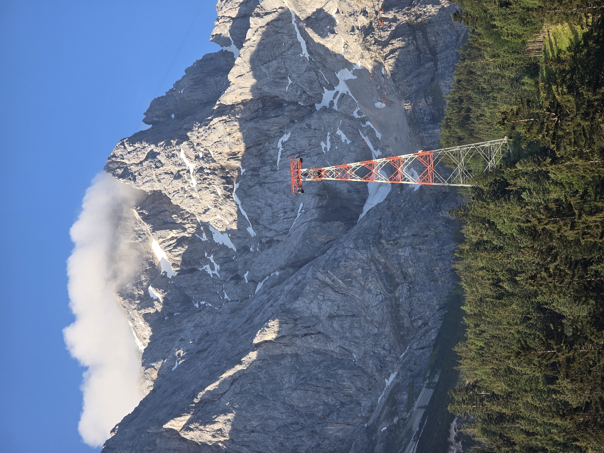 This image depicts a scenic mountain landscape near Grainau, Germany, likely at Zugspitze. A tall, red and white lattice tower, part of a cable car system, stands prominently in the foreground. The tower supports thick cables that stretch across the frame, disappearing towards the mountain peaks. Behind the tower, a dense, dark green forest of coniferous trees blankets the lower slopes. The backdrop is dominated by the imposing, grey, rocky face of a mountain range. Patches of snow are visible on the higher elevations, and a soft, white cloud hovers near the summit of the central peak. The sky is a clear, vibrant blue. There are no people visible in the image. The scene is illuminated by natural daylight, casting shadows that highlight the rugged textures of the mountain and the intricate structure of the tower.