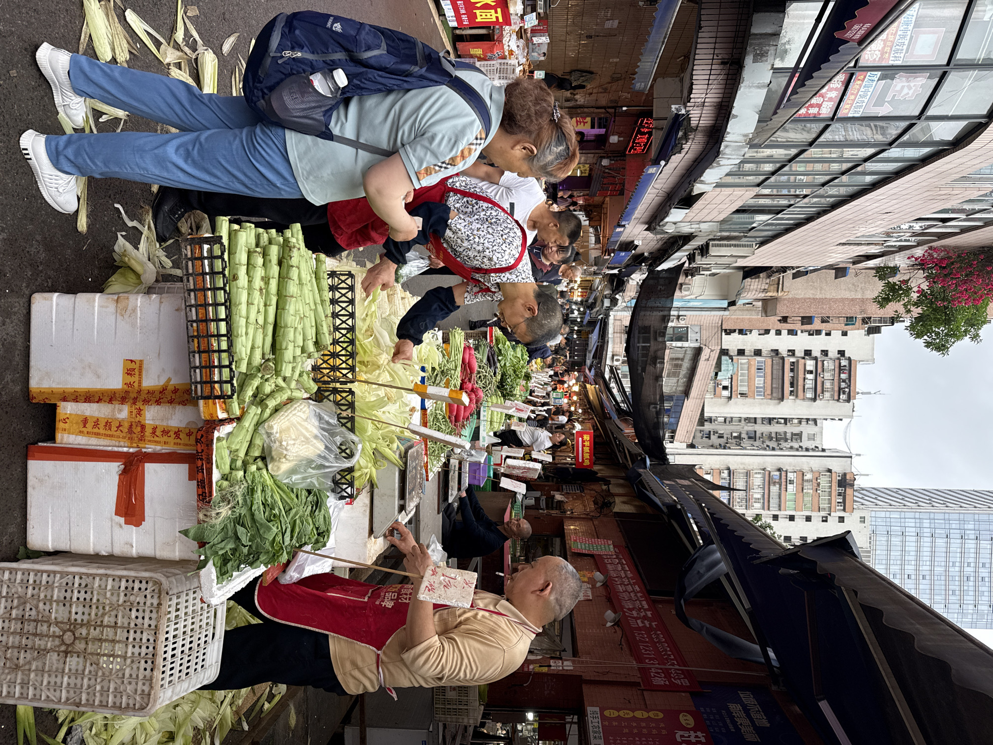 The image captures a bustling street market scene in Nan'an District, China. Numerous individuals are present, predominantly focused on selecting and purchasing vegetables from vendors. A vendor in a red apron appears to be calculating a transaction, using chopsticks as a counting aid. Various vegetables are displayed for sale, including bundles of green stalks, leafy greens, and corn still in their husks. A mixture of older buildings and modern high-rises are visible in the background, reflecting the urban environment. Overhanging canopies and awnings shade portions of the street. Storefront signs and advertisements line the buildings, written in Chinese characters.