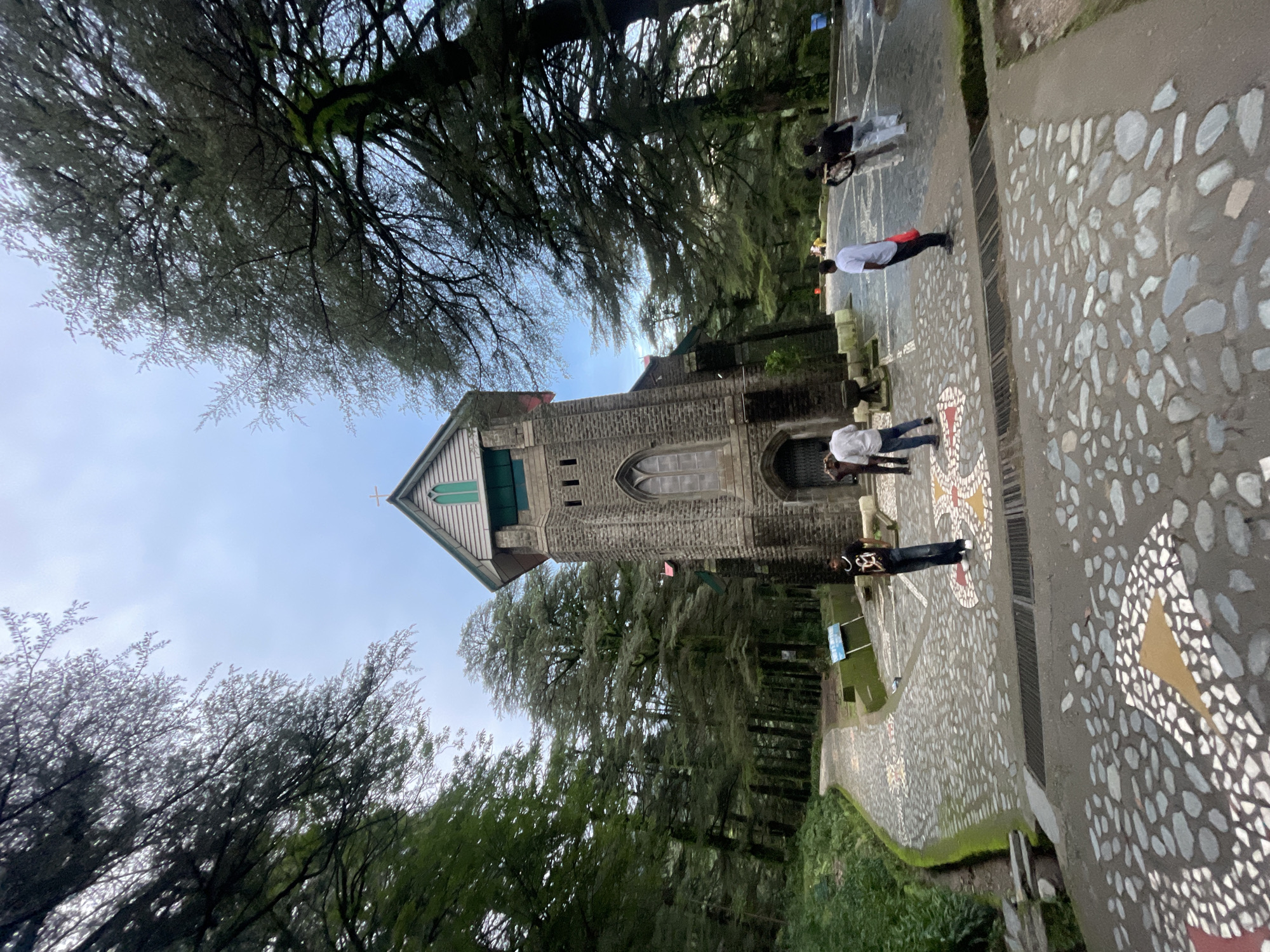 The image shows a stone church in Dharamkot, India, surrounded by tall trees. The church has a tall, narrow tower with a green roof and a cross on top. The facade of the church is made of stone blocks, and it features a tall, arched window. In front of the church, there is a paved walkway with decorative stone patterns. Several people are walking on the walkway, some are standing and looking around. The environment is lush with green trees and foliage, suggesting a natural setting. The sky is overcast with a slightly cloudy appearance.