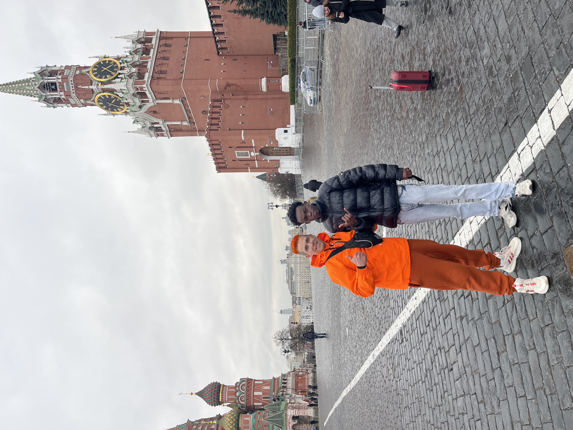 The image depicts two young men standing on a cobblestone surface marked with a white line, likely a pedestrian crossing. One man, wearing an orange tracksuit, is smiling and giving a thumbs-up. The other man, wearing a black puffer jacket and light-colored jeans, is also smiling. The location is identifiable as Red Square in Moscow, Russia. Prominent in the background is the Kremlin's Spasskaya Tower with its distinctive clock face. A section of St. Basil's Cathedral is visible in the lower left. In the background, other people and a red suitcase can be seen. The weather appears overcast.