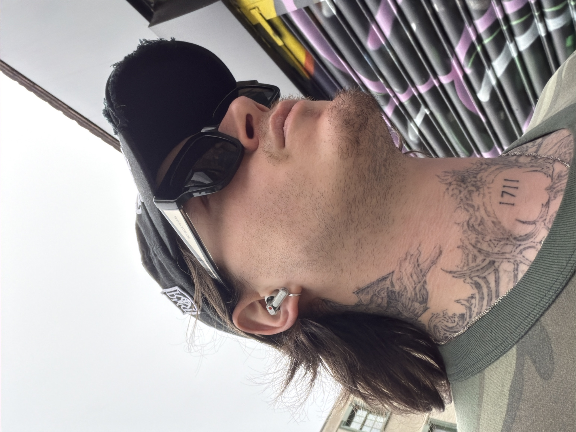 A man with a black baseball cap and sunglasses is photographed from a low angle. He is wearing a camouflage shirt and has tattoos on his neck. The man has an earbud in his ear. He is positioned in front of a colorful graffiti wall. The location is likely in a city setting, possibly London, United Kingdom.
