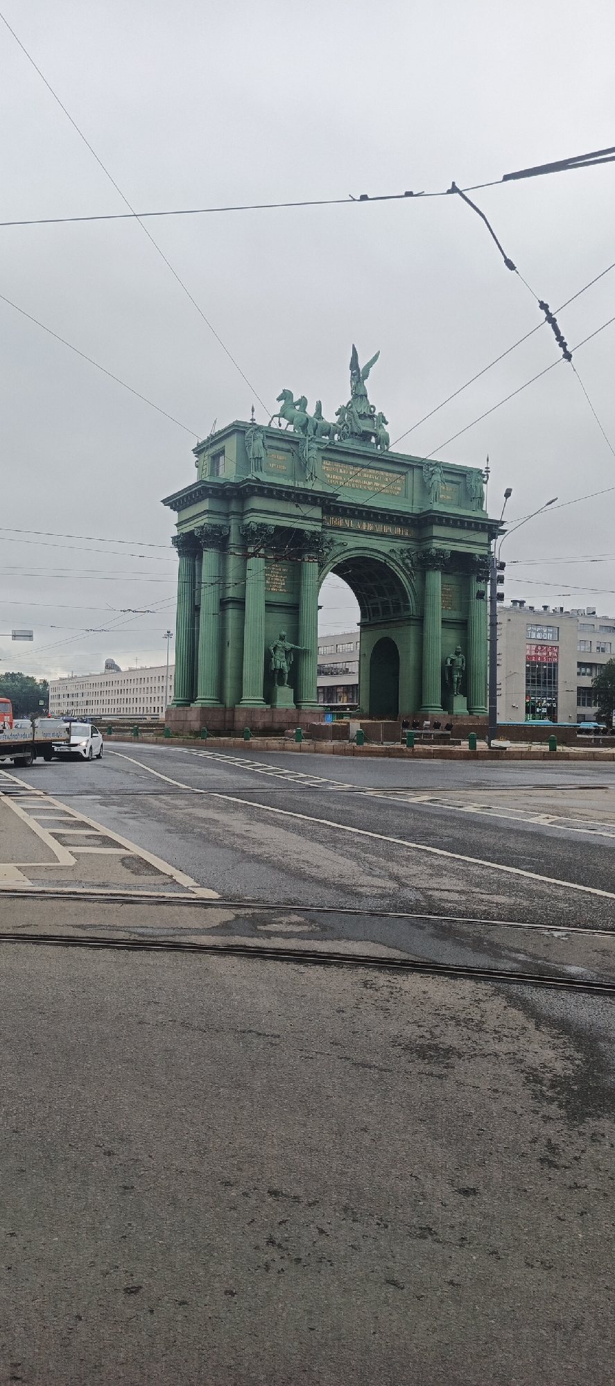 The image depicts a wide-angle, eye-level view of the Narva Triumphal Arch in Saint Petersburg, Russia. The arch, an imposing structure with a distinct green patina, stands prominently in the center of the frame. It is adorned with sculptures of horses and chariots at its apex, as well as figures of soldiers on its sides. The arch is set against a backdrop of an overcast sky and a street scene.
In the foreground, a paved road with tram tracks is visible, appearing damp and reflecting the muted light. White dashed lines mark the lanes of traffic. To the left of the arch, a white car is seen driving away from the camera, and further left, a truck with a trailer is partially visible. In the background, behind the arch, a long, white building with numerous windows stretches across the horizon. Other buildings, some with signage, are also present on either side of the arch. Overhead, power lines are strung across the sky, supported by poles. The overall atmosphere is somber due to the cloudy weather. No people are clearly identifiable in the image.