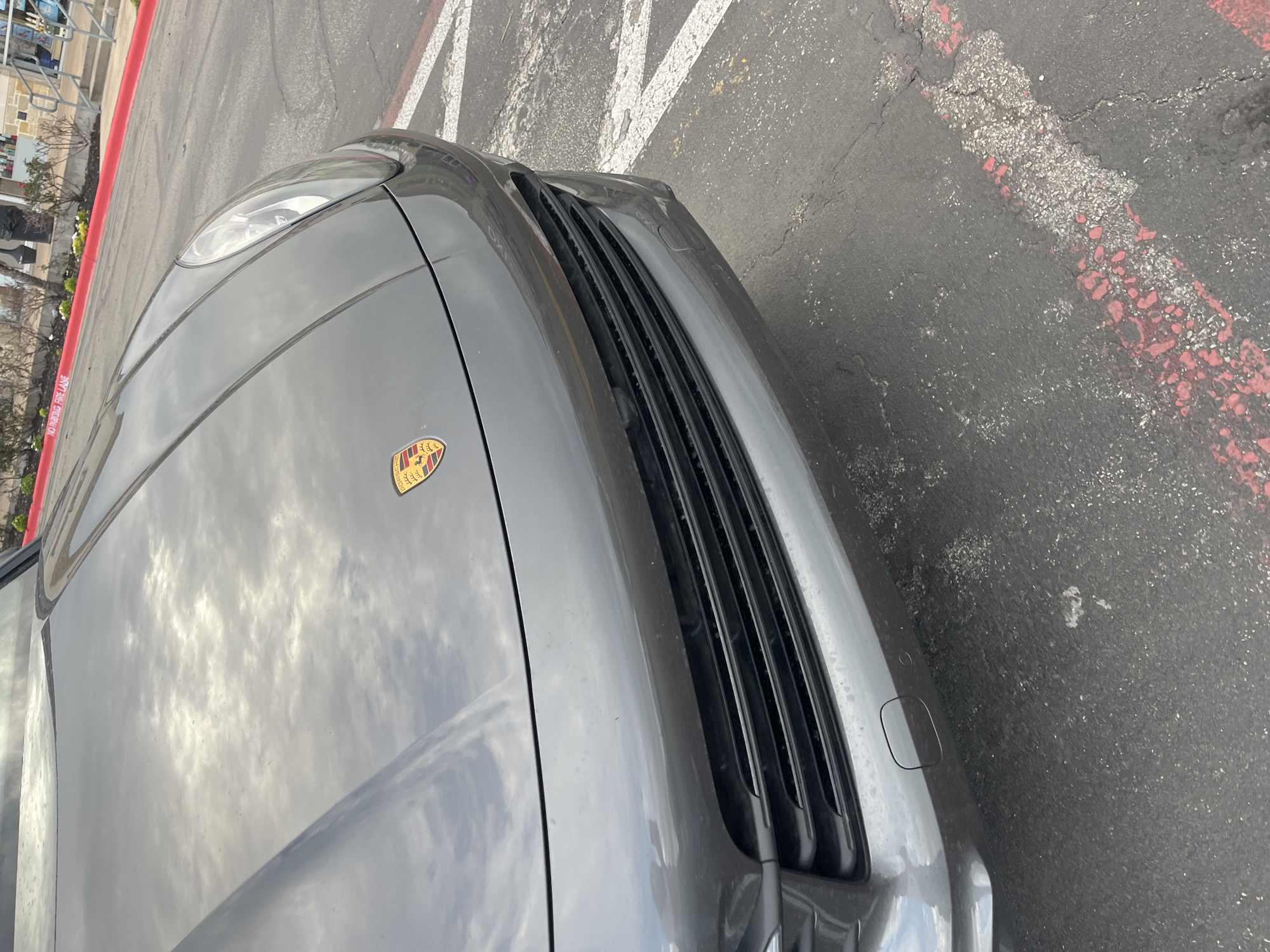 The image shows the front of a gray Porsche parked on an asphalt surface with faded white and red painted lines. The Porsche emblem is visible on the hood, and a reflection of the sky is present on the car's surface. A red curb is visible on the left edge of the image. Based on visual clues of the surroundings of the original image, it is probable the location is in the city of San Marcos, United States of America.