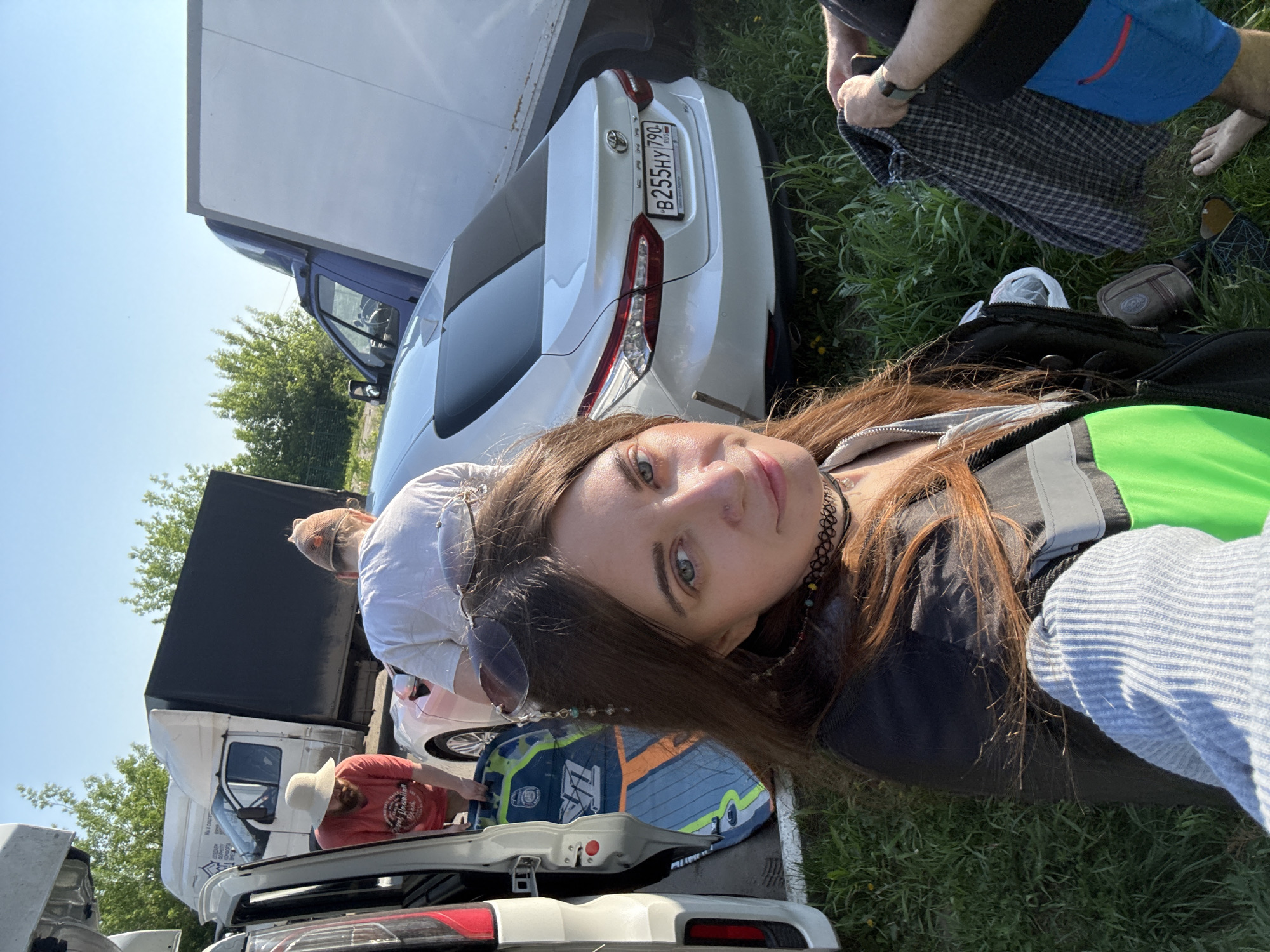 A photograph captures a young woman with long, brown hair taking a selfie. She is wearing a black choker necklace, a green shirt, and a gray jacket. Several individuals are visible in the background. A man wearing a red shirt and a hat is seen standing near the opened trunk of a white car. Another man, shirtless, is wearing blue shorts and a checkered shirt. Cars surround the group, including a white sedan with a Russian license plate (B255HY 790). In the background is also a large truck with a dark cargo container. The scene takes place in a grassy area under bright sunlight, suggesting it is outdoors. The location is likely in Dzerzhinsky, Russia.