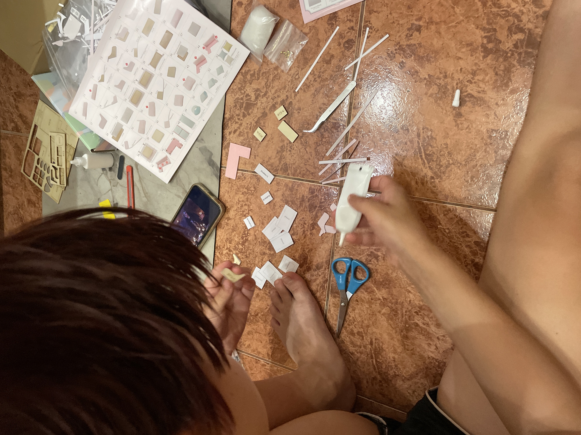 A person is sitting on the floor, assembling a miniature model. The person has dark hair and is wearing black shorts. They are holding small pieces of paper and a white object, possibly a tool or another part of the model. In front of them are scattered pieces of the model, glue, scissors, tweezers, and instructions. The floor is tiled with a terracotta color. A mobile phone is visible next to the instructions. The scene appears to be indoors, with the lighting suggesting it's daytime. There are no visual cues in the image that indicate the location is in Sochi, Russia.
