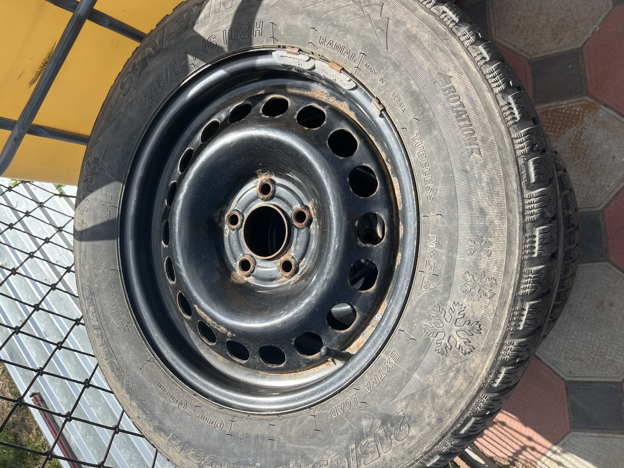 This media file shows a close-up, overhead view of a single tire mounted on a black steel wheel. The tire is a 215/65 R16 102H size and appears to be a winter tire, indicated by the "M+S" marking and snowflake symbols on its sidewall. The tire is branded with "SUN-W" and has text indicating it is "RADIAL" and "TUBELESS." It was "MADE IN SERBIA." The wheel is a standard steel rim with multiple circular cutouts. There are a few small tire balancing weights attached to the rim's edge. The tire shows some signs of wear and dirt.
The tire and wheel assembly is positioned outdoors. In the background, a yellow surface, possibly part of a vehicle or structure, is visible, along with a black metal mesh fence. The ground beneath and around the tire is paved with interlocking concrete tiles in red and gray colors, suggesting an outdoor hard surface like a driveway or patio. The lighting indicates it is daytime, with some shadows cast on the ground, suggesting the sun is at a moderate angle. The overall scene is a still life focusing on the tire and wheel, with no people or overt activity depicted. The context of "Bohunove, Ukraine" provides a geographical location for the image.