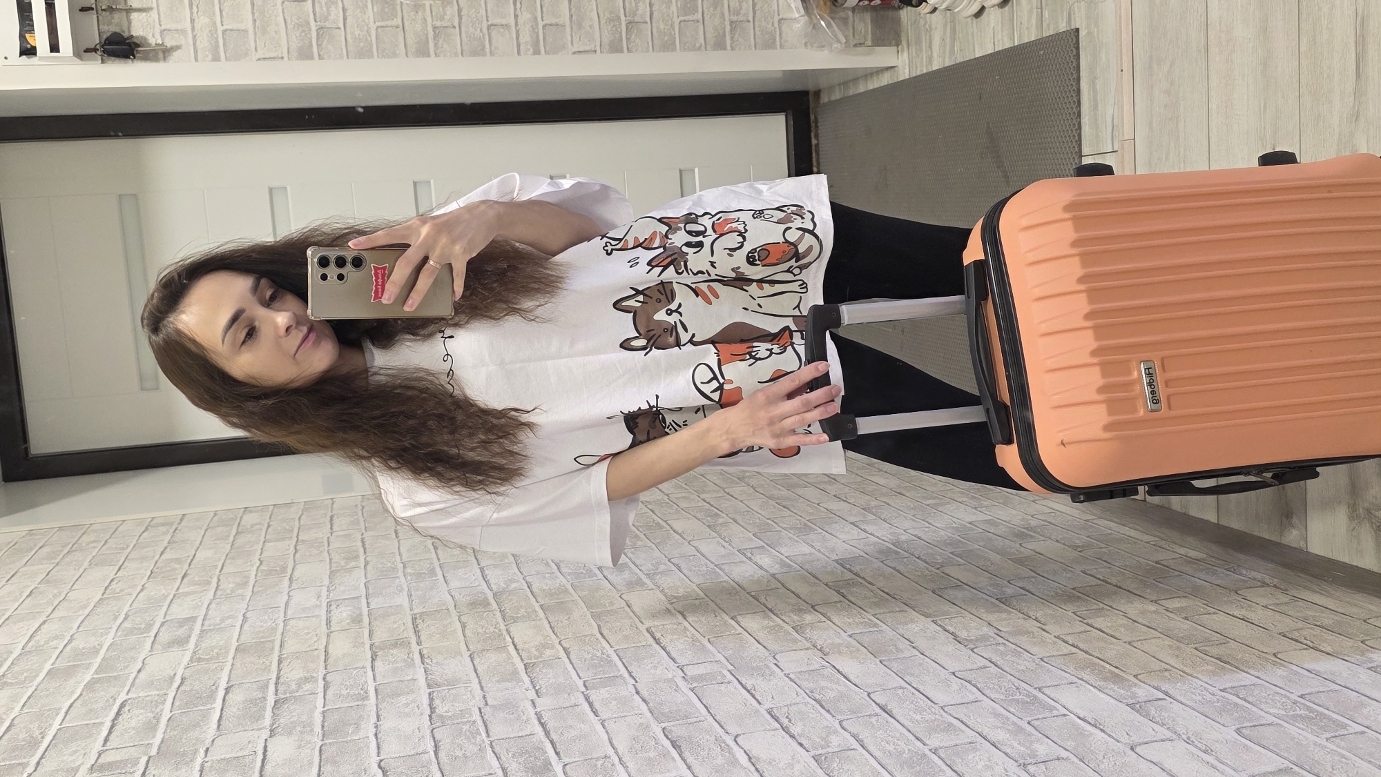 A young woman with long, dark, curly hair stands in a hallway and takes a selfie in a mirror. She is wearing a white t-shirt with a cartoon cat print and black leggings. She is holding a smartphone in her right hand and her left hand rests on the handle of an orange rolling suitcase. The hallway has a white brick-patterned wallpaper on the left wall, and a white door with a black frame is visible in the background. A gray mat is on the floor.