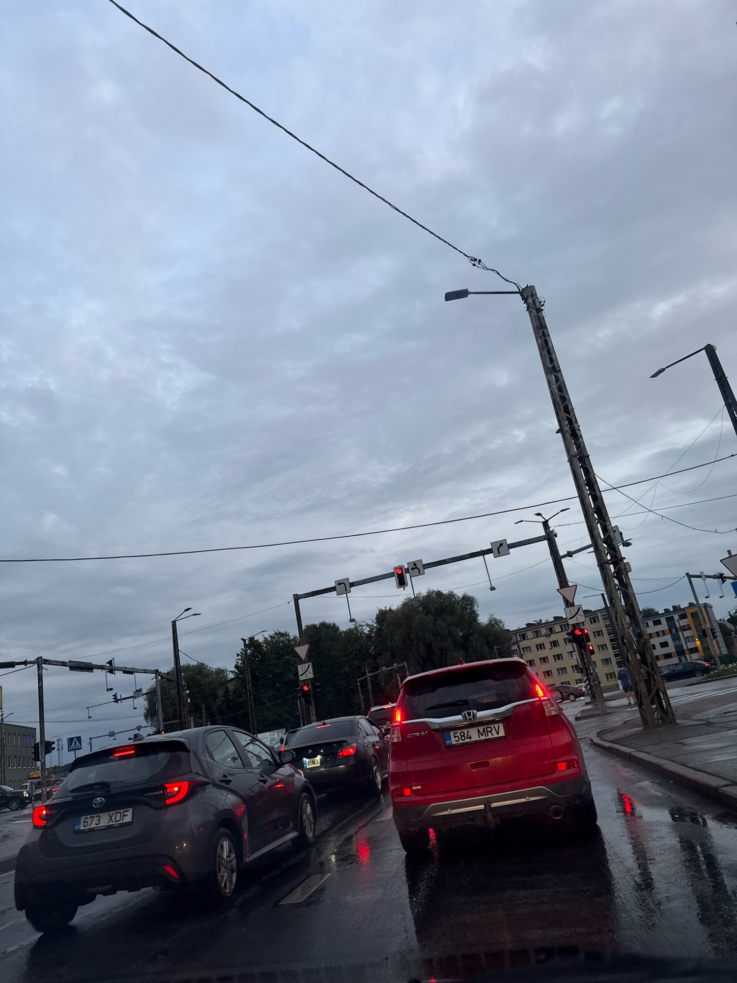 The image depicts a street scene in Tallinn, Estonia, under an overcast sky. Multiple vehicles are stopped at a traffic light, suggesting moderate traffic congestion. The road surface appears wet, indicating recent rain or damp conditions. In the foreground, a dark grey hatchback, identified by its license plate "673 XDF," is prominent. To its right, a red Honda CR-V with license plate "584 MRV" is also visible. Further back, other vehicles and buildings line the street. Traffic lights are visible, with at least one showing a red signal. Utility poles with wires and streetlights are also part of the urban infrastructure. The overall atmosphere is that of a typical city street during a cloudy day.