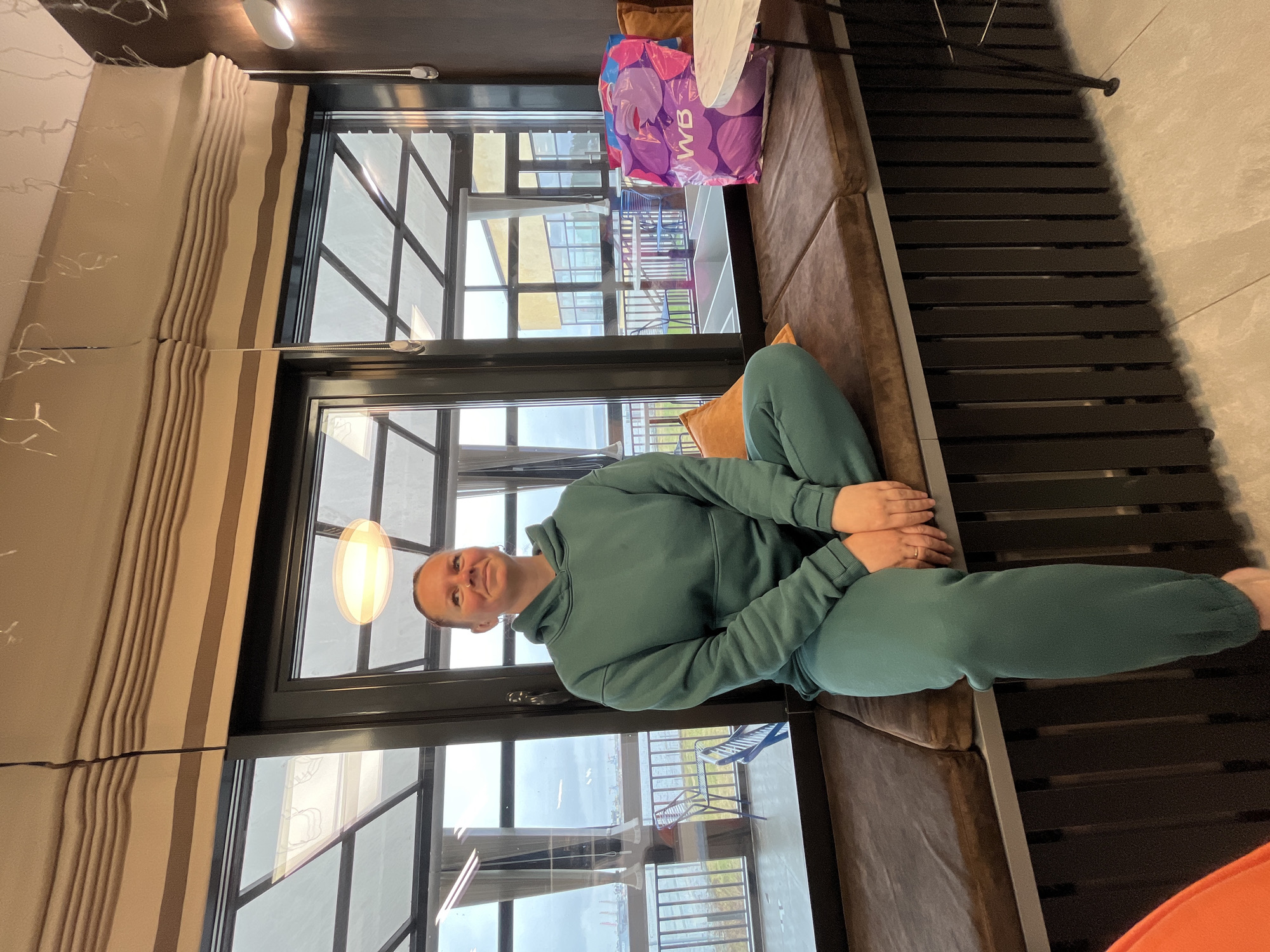 The image depicts a woman seated comfortably on a plush brown sofa or bench, looking towards the camera with a gentle smile. She is wearing a teal or dark green hooded tracksuit, with her hands clasped in her lap and her hair pulled back. The setting is an indoor space in Chaikovsky, Russia, with a large window providing an expansive view of the outdoors.
The window features dark framing and multiple panes, reflecting a contemporary design. Through the window, a body of water, possibly a river or reservoir, extends into the distance, with faint outlines of land or structures on the horizon. An outdoor balcony or terrace is visible directly outside the window, furnished with what appears to be blue and white chairs. Beige Roman blinds with vertical stripes are partially drawn on the left side of the window, and a light fixture with an oval diffuser hangs nearby.
To the woman's right on the seating area, a purple shopping bag with the white letters "WB" (likely indicating Wildberries) is placed next to a small, round, white table surface. Adjacent to the sofa, a dark slatted structure, possibly a radiator cover or floor grating, runs along the floor. A hint of an orange cushion is also visible in the bottom right corner of the frame. The bright natural light streaming through the window suggests it is daytime with clear weather. The scene conveys a sense of peaceful relaxation in a modern interior with a scenic outdoor vista.