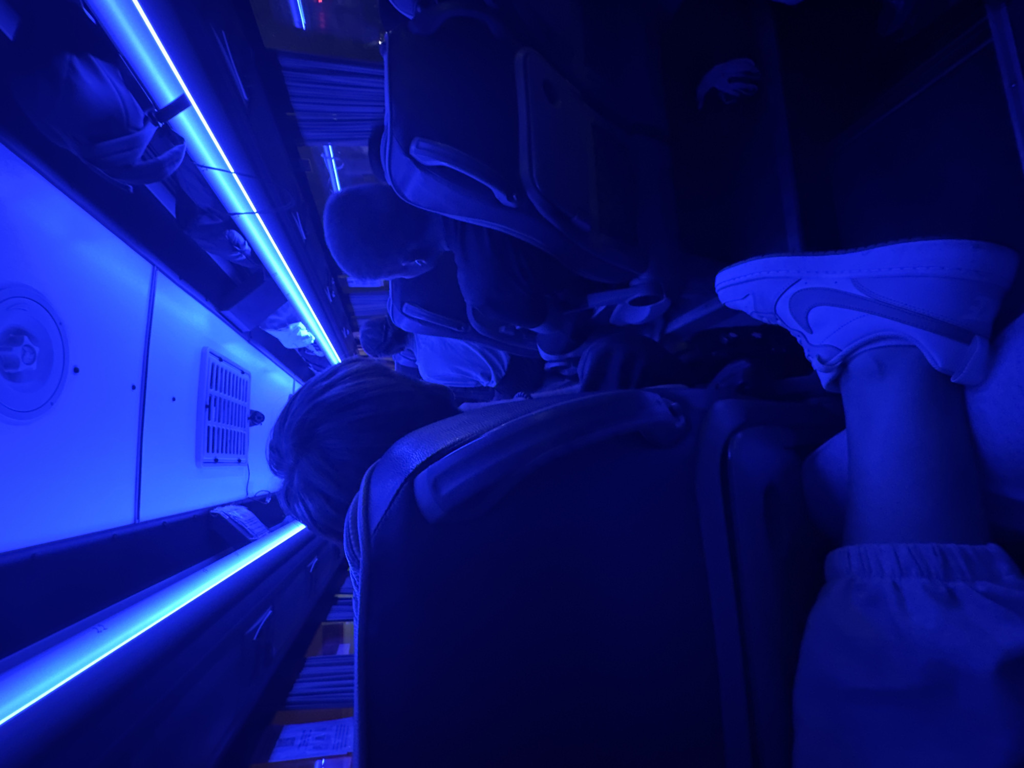 The image depicts the interior of a dimly lit bus or passenger van, illuminated by bright blue LED strip lights running along the ceiling. Several passengers are visible, seated facing forward in rows of dark-colored seats. The perspective is from a passenger in the foreground, with their leg and white sneaker prominently displayed, crossed over another seat. The overall ambiance is dark and atmospheric due to the blue lighting, suggesting nighttime or an enclosed space. There is no indication of the city of Staroe Abdulovo, Russia, or any specific urban setting.