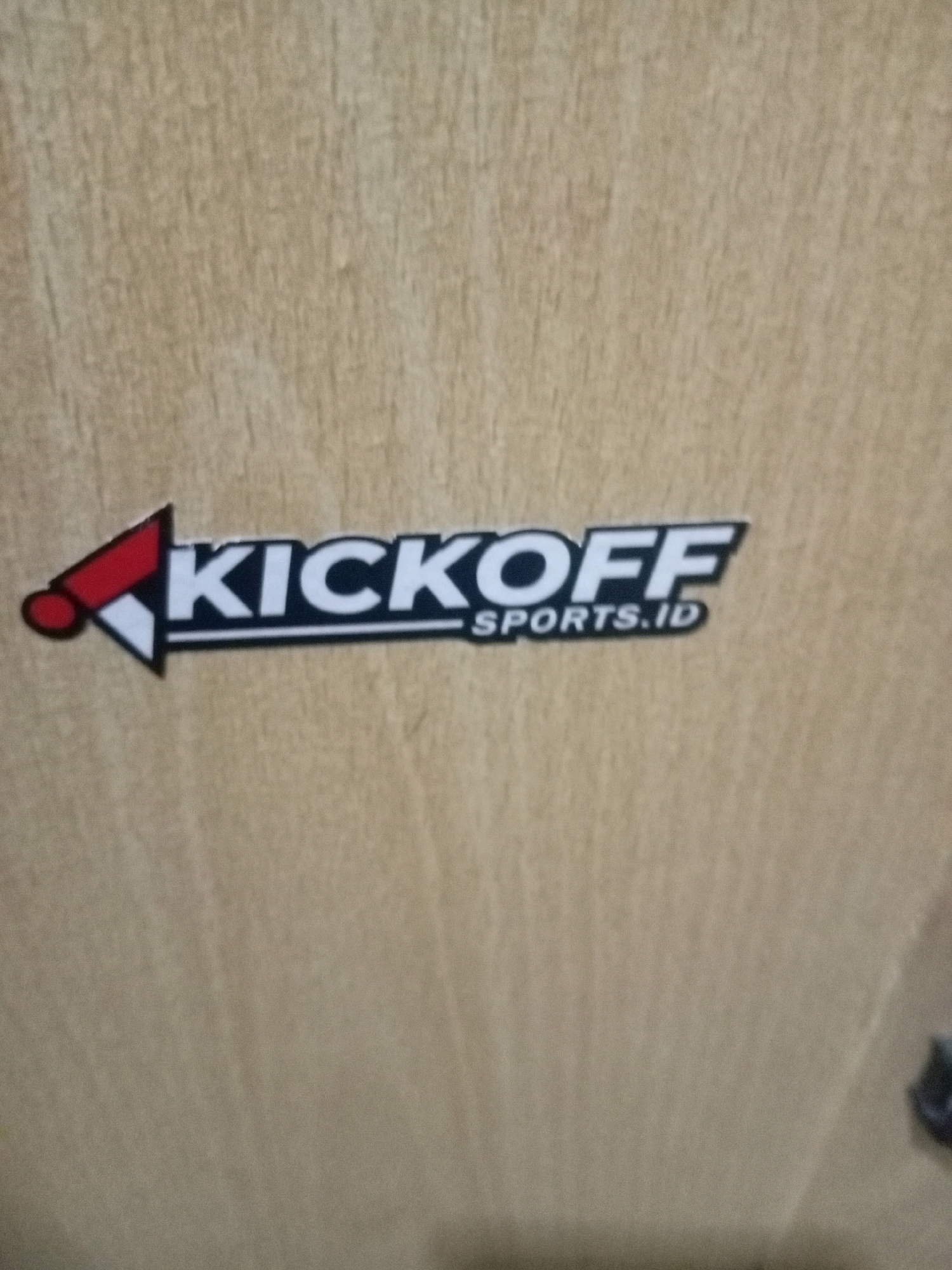 The image shows a close-up of a logo on a light-colored, textured surface, likely wood or laminate. The logo features the word "KICKOFF" in large, bold, white capital letters outlined in black. Below "KICKOFF," in smaller white capital letters, is "SPORTS.ID." To the left of the text is a graphic element resembling an arrow pointing left. This arrow is red at the tip and white towards the text, with a black outline. The background is out of focus, displaying a uniform, light brown color with subtle vertical grain patterns. The lighting is consistent and appears to be from above or the front, with no harsh shadows. The scene is devoid of any people or specific environmental features that would indicate a city location or Batam, Indonesia.