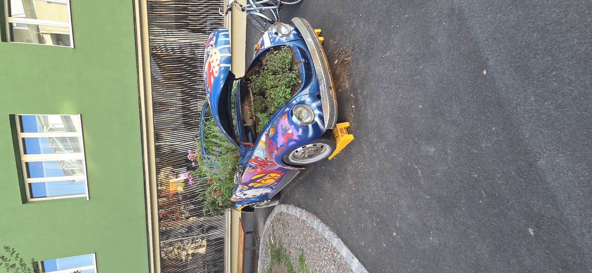 The image depicts a vibrant, custom-painted blue Volkswagen Beetle, likely a convertible, repurposed as a planter. The car is adorned with colorful graffiti-style art, including red, yellow, and white abstract designs and figures. The front hood is open, revealing a lush planting of green foliage and various flowering plants, including pink and purple blooms. The car rests on bright yellow wheel chocks.
The setting appears to be an urban environment. To the left, a wooden bench sits beside a gravel pathway that curves away from the car. In the background, a building with a distinctive green facade and large windows is visible. Below the windows is a long, horizontally striped section, possibly part of the building's exterior or a decorative element. The ground around the car is paved with asphalt. A bicycle is partially visible to the right of the car, leaning on something.
Based on the context provided, this scene is located in Bregenz, Austria. The specific location appears to be an outdoor public space, possibly a pedestrian area or courtyard adjacent to a building. The time of day is likely daytime, judging by the lighting and reflections in the windows. There are no people visible in the image.