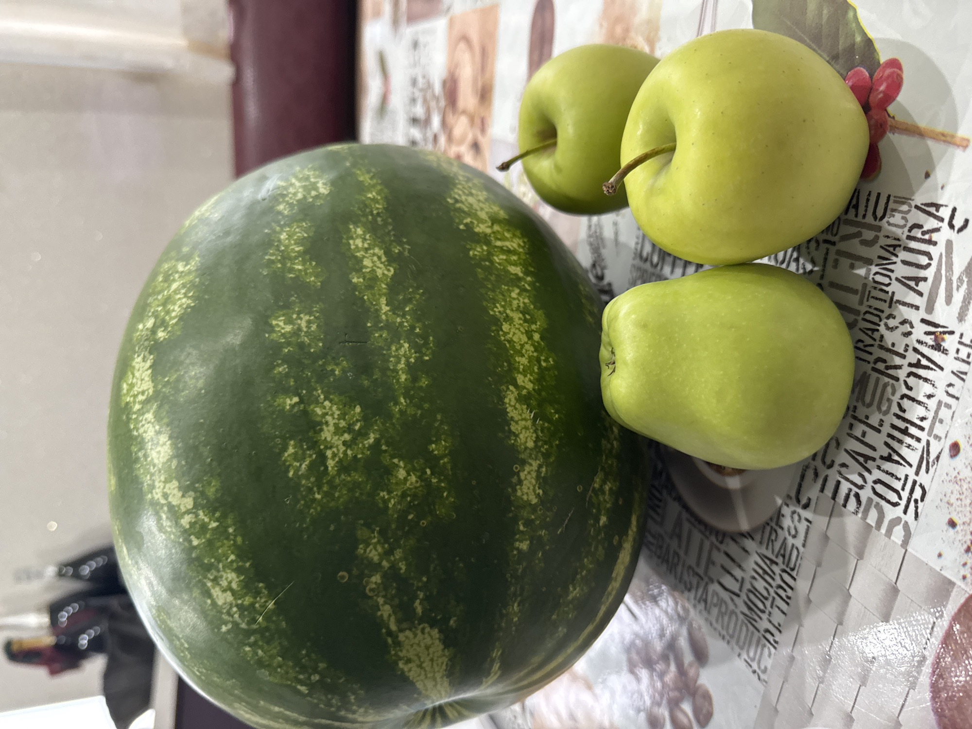 A large, green, striped watermelon sits on a table covered with a tablecloth featuring a pattern of coffee-related text and images. Three green apples are arranged in front of the watermelon. The lighting suggests an indoor setting, possibly a kitchen or dining area. In the blurred background, there are some bottles and a dark red upholstered surface. The image appears to be taken at eye level, looking slightly down at the fruits. The overall composition focuses on the arrangement of the fruits on the patterned tablecloth.