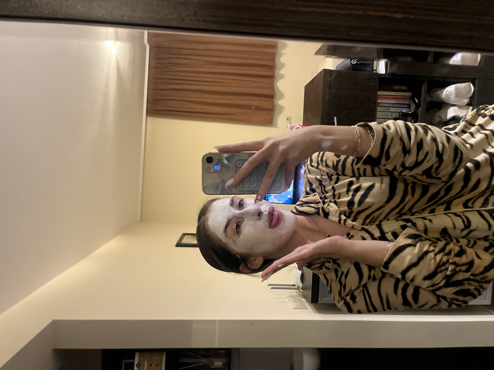 A young woman in a tiger-striped pajama set is taking a selfie in a mirror. She has a white facial mask on. The background shows a room interior, including a dresser, a window with blinds, and a bookshelf. It is not possible to ascertain the location of the scene as Sofia, Bulgaria without further information.