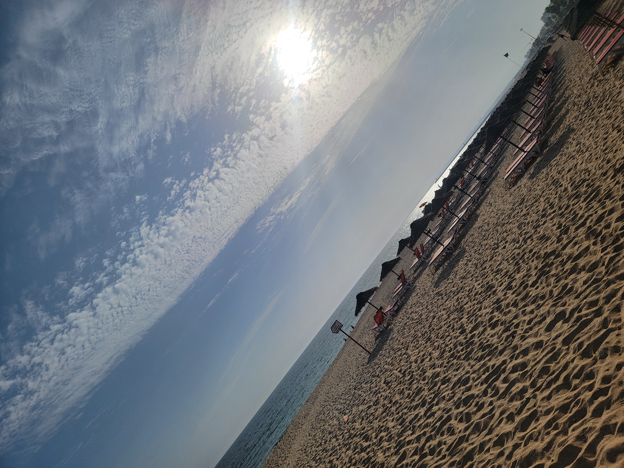 This is a diagonal shot of a beach in Quarteira, Portugal, likely during the daytime. The scene is characterized by a vast expanse of sand, meticulously patterned with ripples from the wind or receding tides. A long row of empty beach chairs and umbrellas stretches along the coastline, suggesting a quiet moment before or after peak visitor hours. The sea is visible to the left, its surface shimmering under the bright sun, which is prominently featured in the upper left quadrant of the image. The sky is a clear blue, dotted with scattered, wispy white clouds that catch the sunlight. In the background, along the edge of the beach, a line of buildings can be seen, hinting at the urban proximity of this coastal area. The overall mood is tranquil and inviting, evoking a sense of relaxation and summer vacation. A small signpost with some text is visible on the beach between the chairs and the water.
