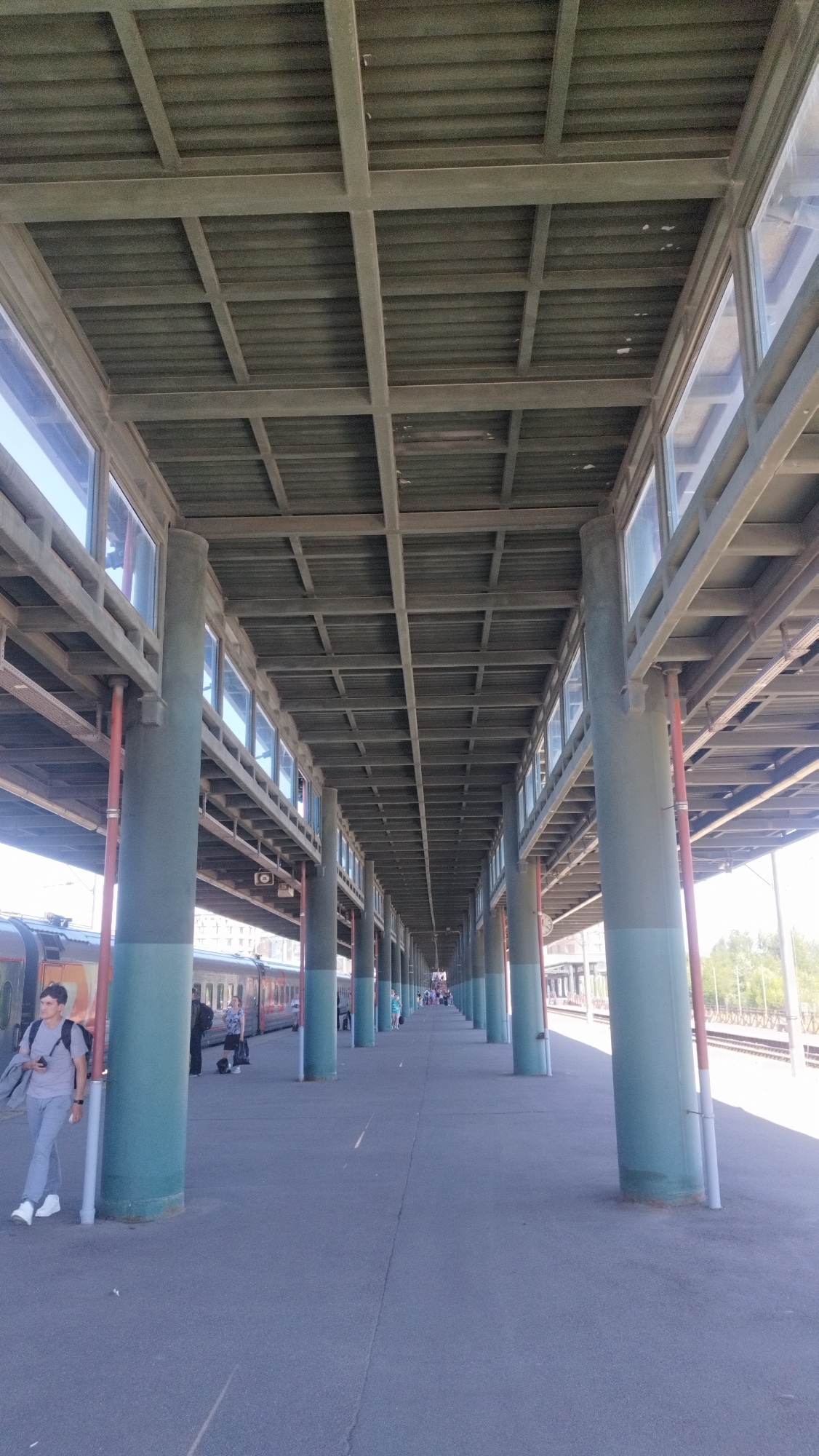 An outdoor, low-angle, eye-level, vertical, full shot shows a train station platform with a covered walkway supported by numerous teal-colored cylindrical pillars. The walkway has a metal roof structure with a grid of beams and corrugated panels, and rectangular windows along its length. A train with orange and white livery is stopped at the platform on the left. Several people are visible on the platform, including a young man in a gray t-shirt and light-colored pants standing near a pillar on the left, holding a phone and a bag. Other individuals, appearing as silhouettes or small figures, are dispersed further down the platform. The ground is paved with dark asphalt. The scene is bathed in sunlight, casting shadows and indicating a daytime setting. The location is identified as a train station in an unknown city in Russia.