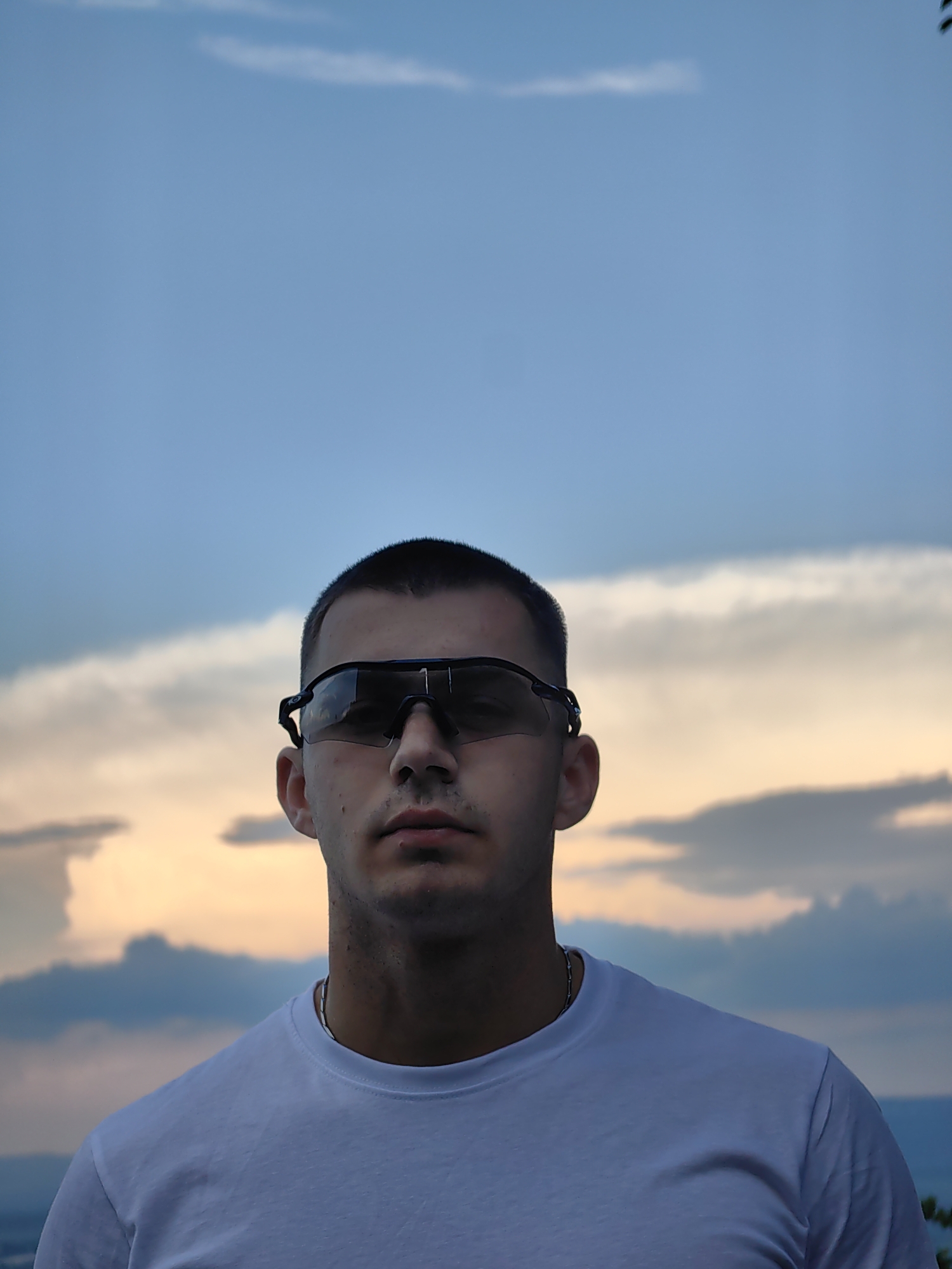 A medium shot depicts a man wearing dark sunglasses and a white t-shirt. He stands against a backdrop of a cloudy sky with tinges of orange and blue, suggesting either sunrise or sunset. The location appears to be outdoors, possibly a slightly elevated position overlooking a landscape or cityscape. The background features the sky and clouds, indicating the subject is in a scenic location. Due to the absence of recognizable landmarks, the location in Shumen, Bulgaria cannot be precisely pinpointed.