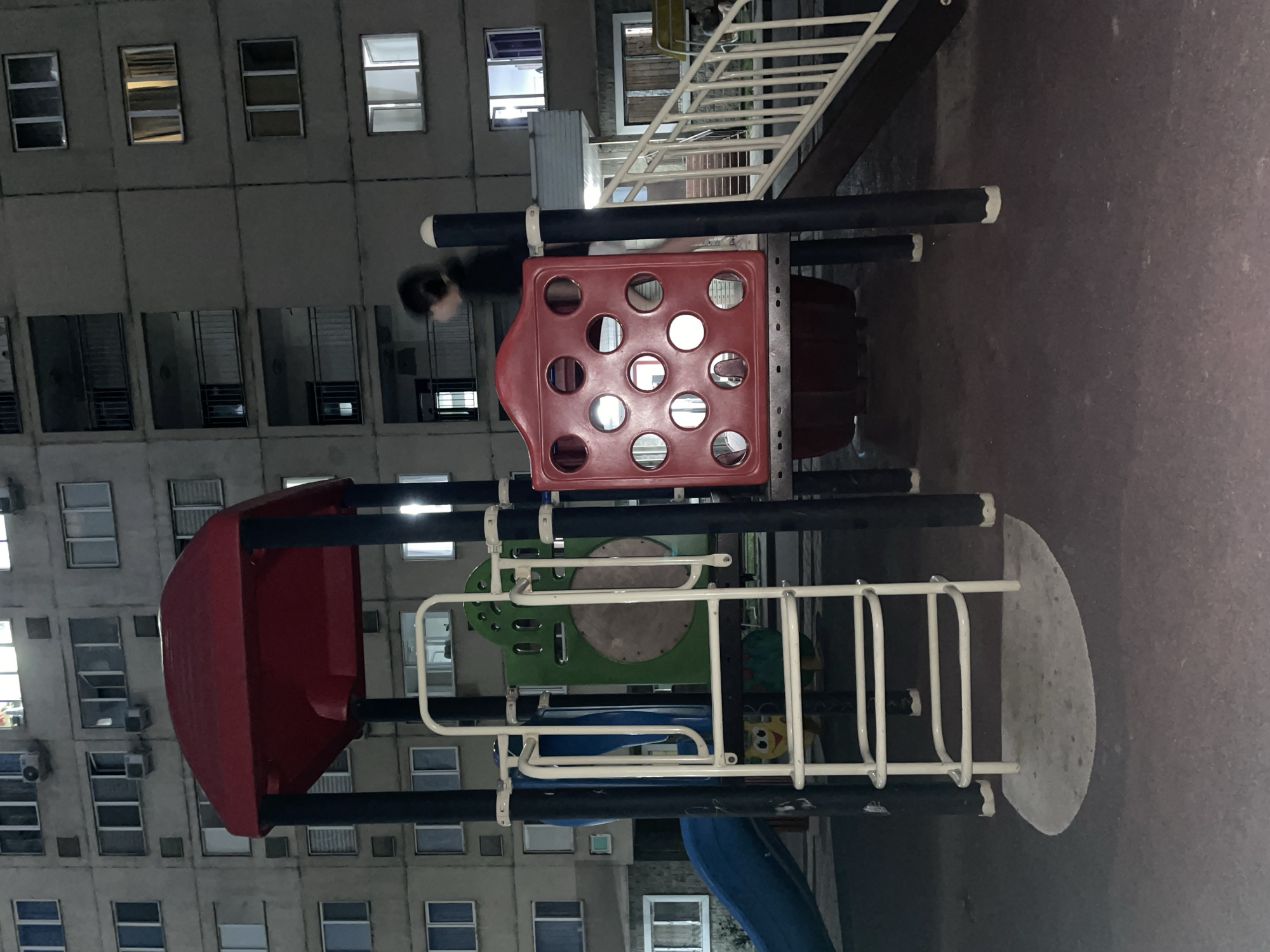 In Almaty, Kazakhstan, at nighttime, a dimly lit playground area is visible in front of a multi-story apartment building. The playground features a red and black play structure with a slide, a ladder, and a red panel with circular cutouts. A person, appearing as a blurred silhouette, is seen moving across the play structure. The apartment building behind the playground has numerous windows, some of which are illuminated. The ground is paved with a dark material.