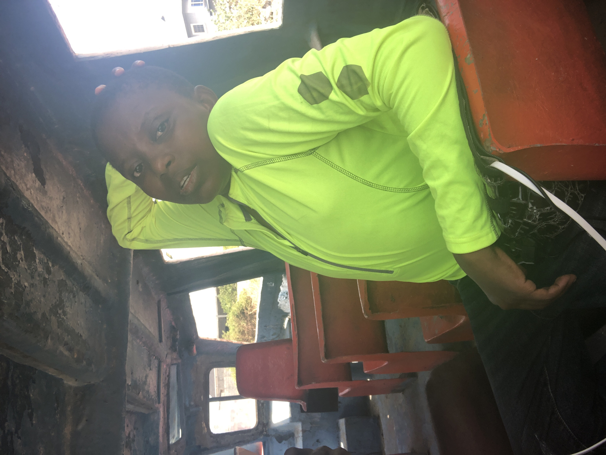 A person wearing a neon green long-sleeved shirt is inside a rusted metal structure with red seating. The interior appears to be part of a derelict vehicle, possibly a train car or similar transport. The lighting is uneven with bright sunlight coming through openings in the structure. Outside, there is a glimpse of a building and trees, suggesting an outdoor location. The exact city is unknown.