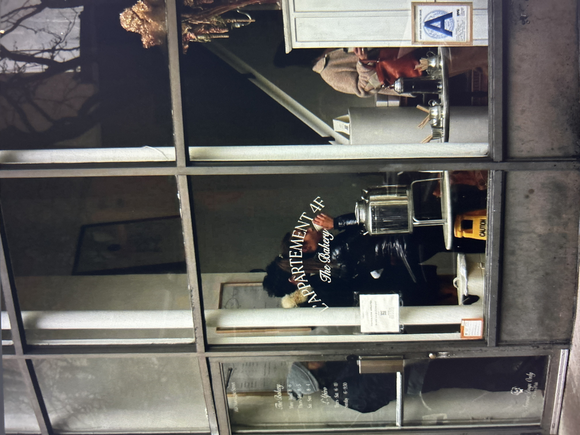 The image shows the exterior of a bakery with large glass windows. Inside, two individuals are visible. One, wearing a grey beanie and a dark jacket, appears to be a customer or staff member looking out through the window. Another person, dressed in black leather, is holding a cup and seemingly interacting with someone else obscured by the reflection. A third person in a tan coat is standing behind a counter on the right side of the image, holding a red bag.
Signs visible on the window include "The Bakery," hours of operation, and "L'APPARTEMENT 4F." A framed health inspection grade with an "A" is displayed on the right. Various objects such as coffee makers, pots, and a yellow caution sign are also present. The environment suggests an urban setting, possibly a street-level storefront.
Based on the provided information, the location of Novaya Usman', Russia, is not ascertainable from the image itself, as there are no visual cues to support this specific geographic designation. The visual content is limited to the interior and exterior of the bakery.