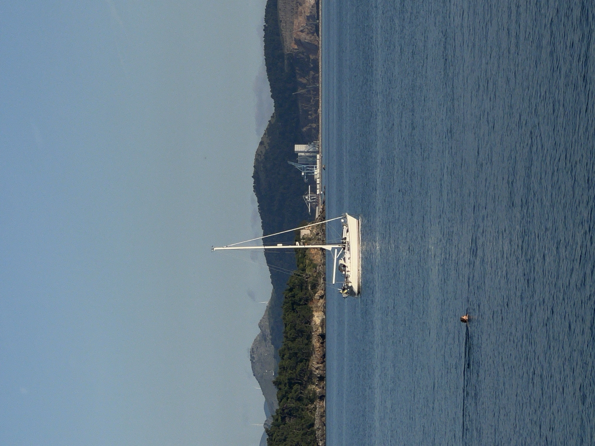 The scene is a vertical view of a marine environment near Sutomore, Montenegro. A white sailboat is in the center of the image, positioned close to a forested coastline with visible mountain ridges in the background. An industrial-looking facility is located on the coast in the background. A single person is swimming or floating in the water, slightly distanced from the boat. The sky is clear.