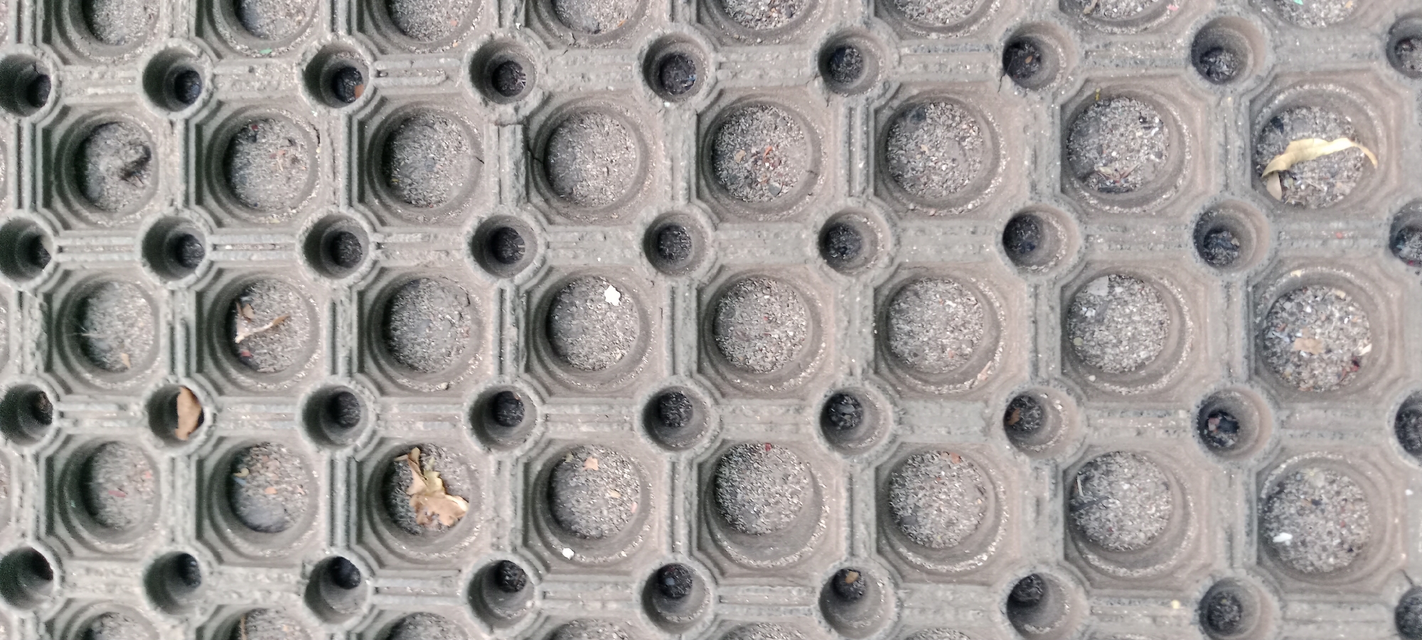 The image displays a close-up, overhead view of a textured, gray rubber mat. The mat has a repeating pattern of raised octagonal shapes, each containing a circular indentation filled with small pebbles and debris. In between these larger indentations are smaller circular holes that appear darker, likely due to shadow or accumulated dirt. There are a few scattered pieces of dried organic matter, such as leaves, within some of the indentations. The mat shows signs of wear and tear, with some of the raised surfaces appearing slightly worn down. The scene is devoid of any human presence or activity.
This image is not specific to any location in Tuban, Indonesia, and provides no contextual information to place it within that city. It is a generic close-up of a textured surface.