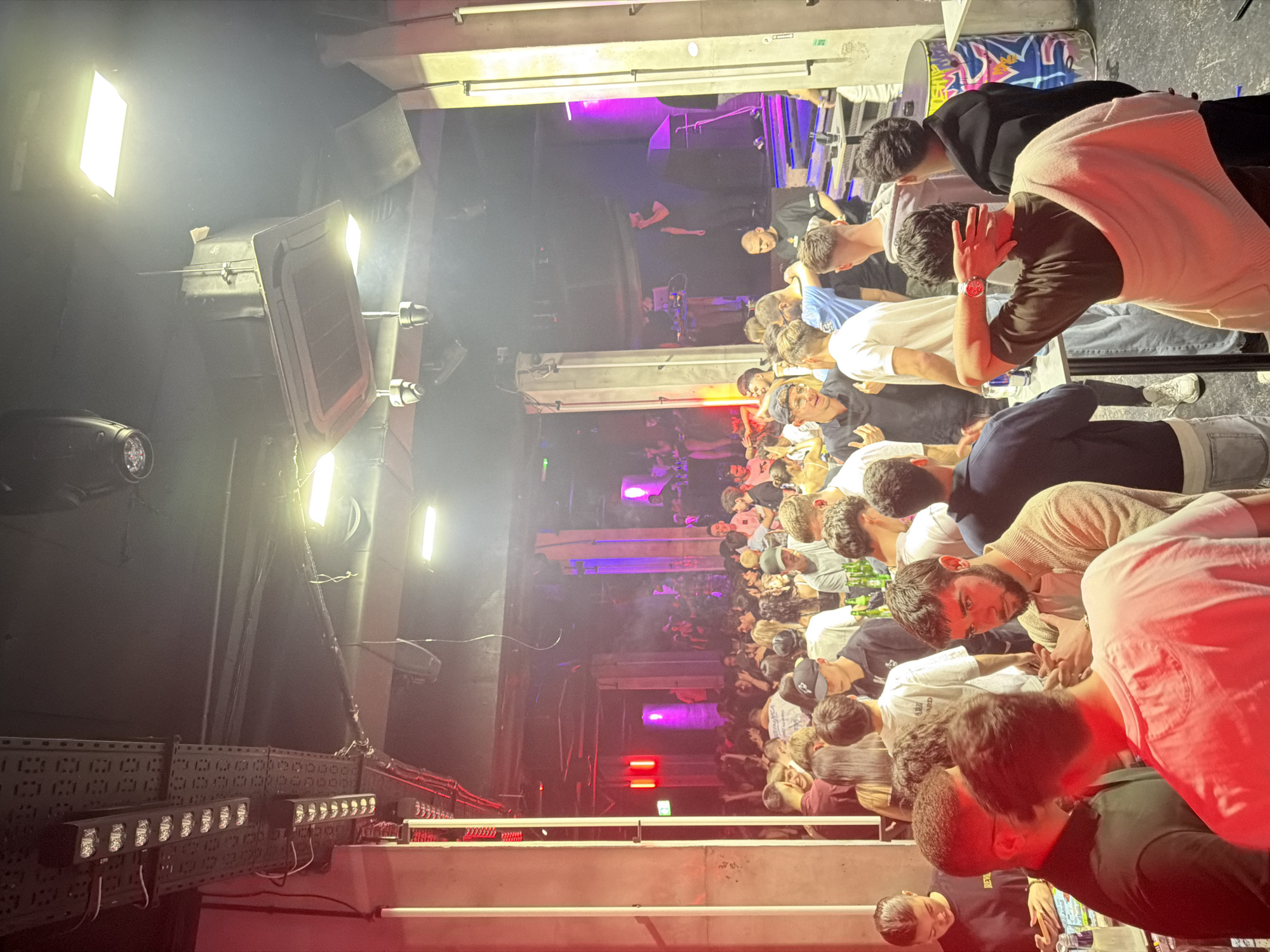 The image depicts a crowded indoor scene, likely a nightclub or music venue, in Varna, Bulgaria. A dense gathering of people, mostly young adults, is observed. The lighting is a mix of bright white spotlights and colored stage lighting, predominantly red and purple. There's a stage area visible in the background, where a DJ or performer appears to be present. The environment seems enclosed, with industrial-style fixtures and elements like metal grates and exposed lighting equipment. The crowd is densely packed, with some people standing close to the camera, others further away. The general atmosphere is energetic and lively, suggesting a social event or entertainment setting.
