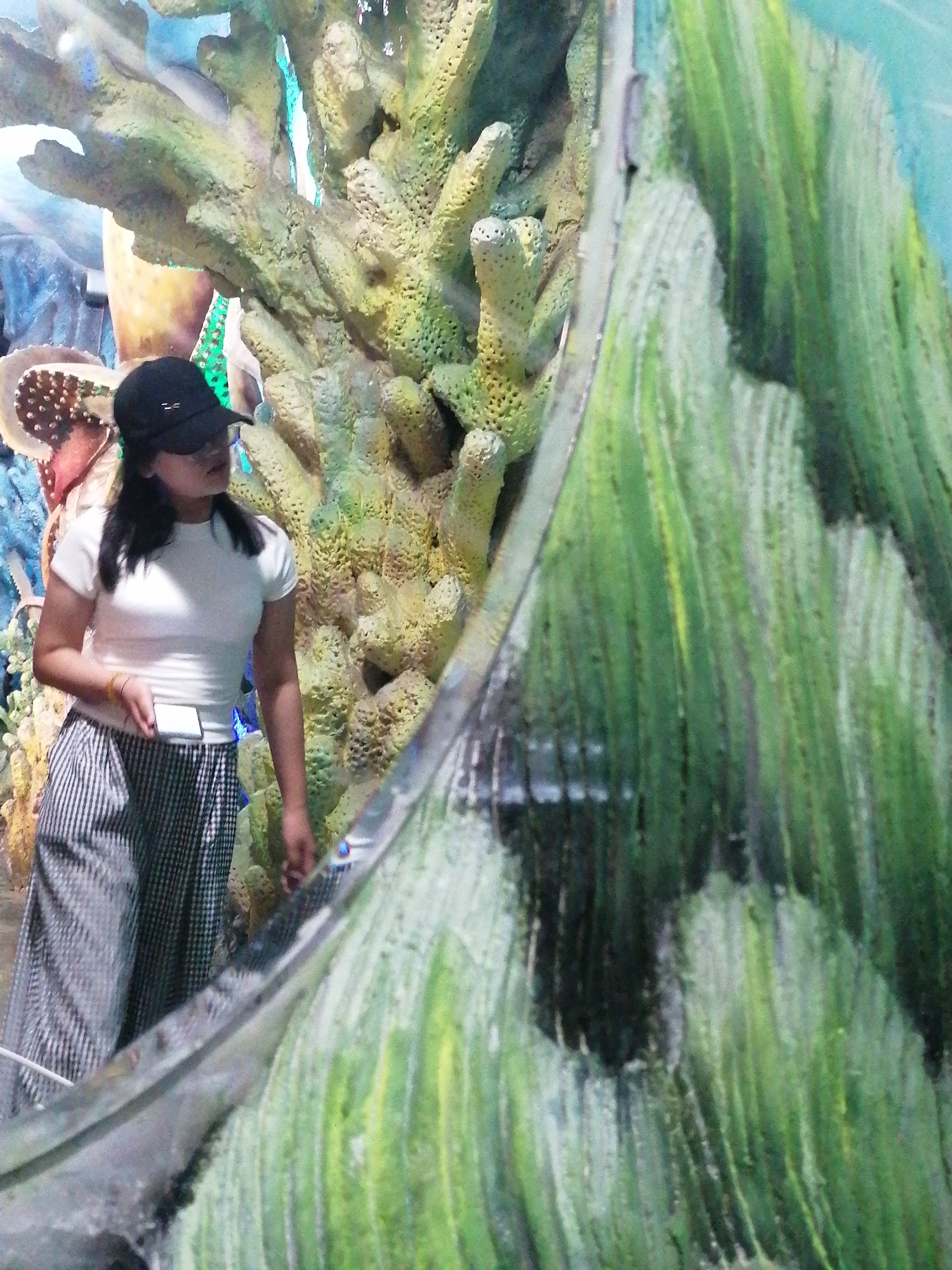 A young woman stands in front of a large aquarium in Da Lat, Vietnam. She wears a white t-shirt, black-and-white striped pants, and a black baseball cap, and holds a white phone. The aquarium behind her contains artificial coral, including one large yellow structure with holey texture to the left and tall green vertical structure to the right. An octopus display is visible in the background.
