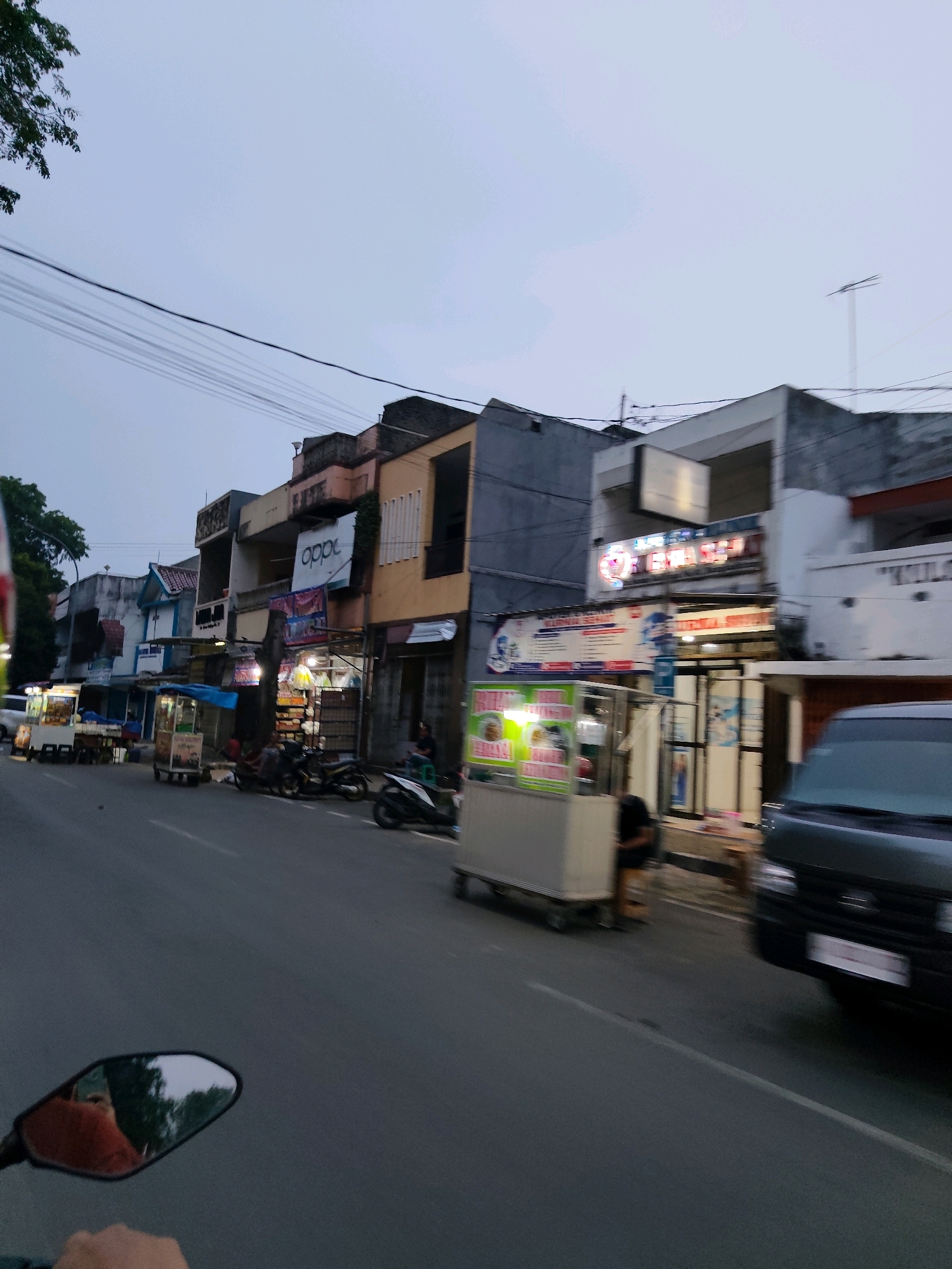 The image depicts a street scene in Rangkasbitung, Indonesia, likely in the late afternoon or early evening as indicated by the dim lighting. The foreground shows a motorcycle's rearview mirror reflecting a person wearing an orange shirt and a glimpse of trees. The street is paved and appears to be moderately trafficked, with several small shops and food stalls lining the right side of the road.
On the right side of the street, there are various commercial establishments, including what appears to be a shop with an "OPPO" sign prominently displayed. Further down the street, there are several food carts or stalls, some with customers or vendors visible. One stall has a white cart with a covered top, and another has a smaller cart with food items displayed. A dark-colored van is parked or moving on the right side of the street.
On the left side of the street, more shops and buildings are visible, some with signage that is difficult to decipher due to distance and image quality. There are also several motorcycles parked along the roadside. The sky above is overcast and light gray, suggesting a cloudy or twilight condition. The overall impression is of a typical urban street in a commercial area of Rangkasbitung.