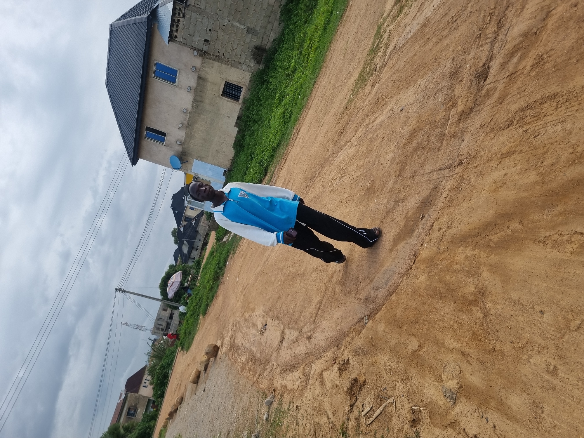 A young Black male, appearing to be in his late teens or early twenties, is walking towards the camera on a dirt road in Jos, Nigeria. He is wearing a blue and white long-sleeved collared shirt and black pants with white stripes. He is looking directly at the camera with a neutral expression. The road is unpaved and appears to be a light brown sandy color, with some tire tracks visible. In the background, there are several buildings, including a two-story light-colored house with a dark gray roof and blue windows. There is also a brick building under construction and some lush green vegetation. The sky is overcast with gray clouds. The overall scene suggests an urban or semi-urban environment.