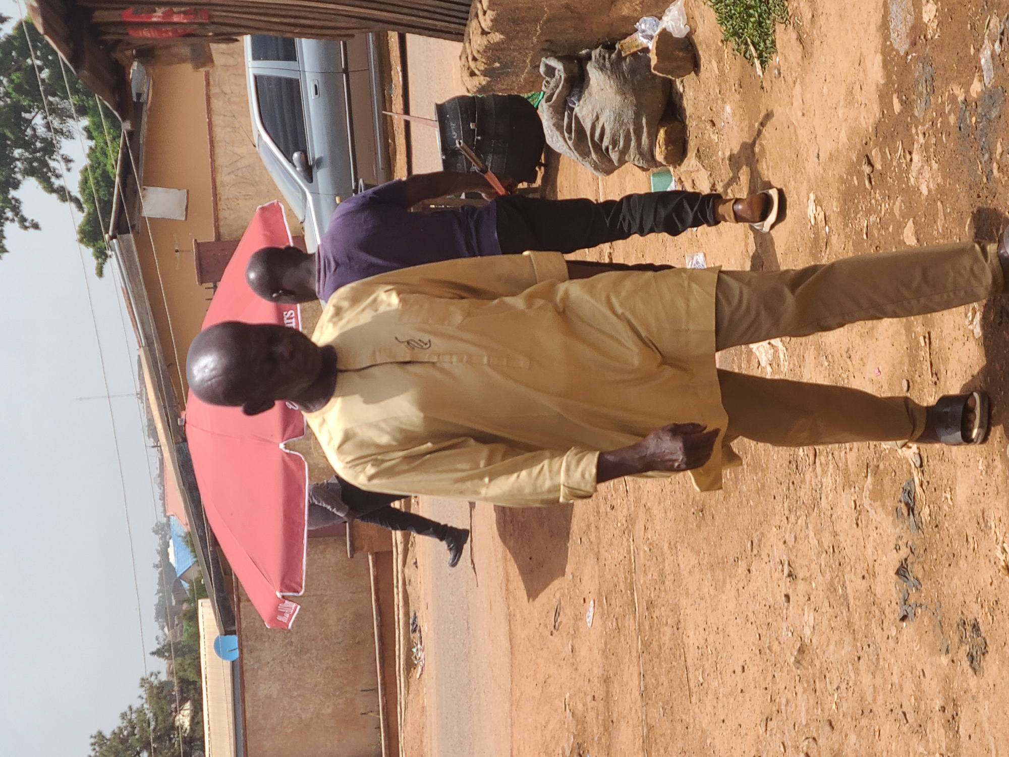 In Jos, Nigeria, the scene captures two individuals on a dirt road, adjacent to a storefront or shaded area with a red umbrella. One man, wearing a light yellow shirt and khaki pants, is in the foreground. Behind him is another man wearing a purple shirt and dark pants is bent over a black bucket. Near the bucket is a gray sack. A silver vehicle is in the background, parked near the storefront. The environment is dusty and appears to be a commercial area.