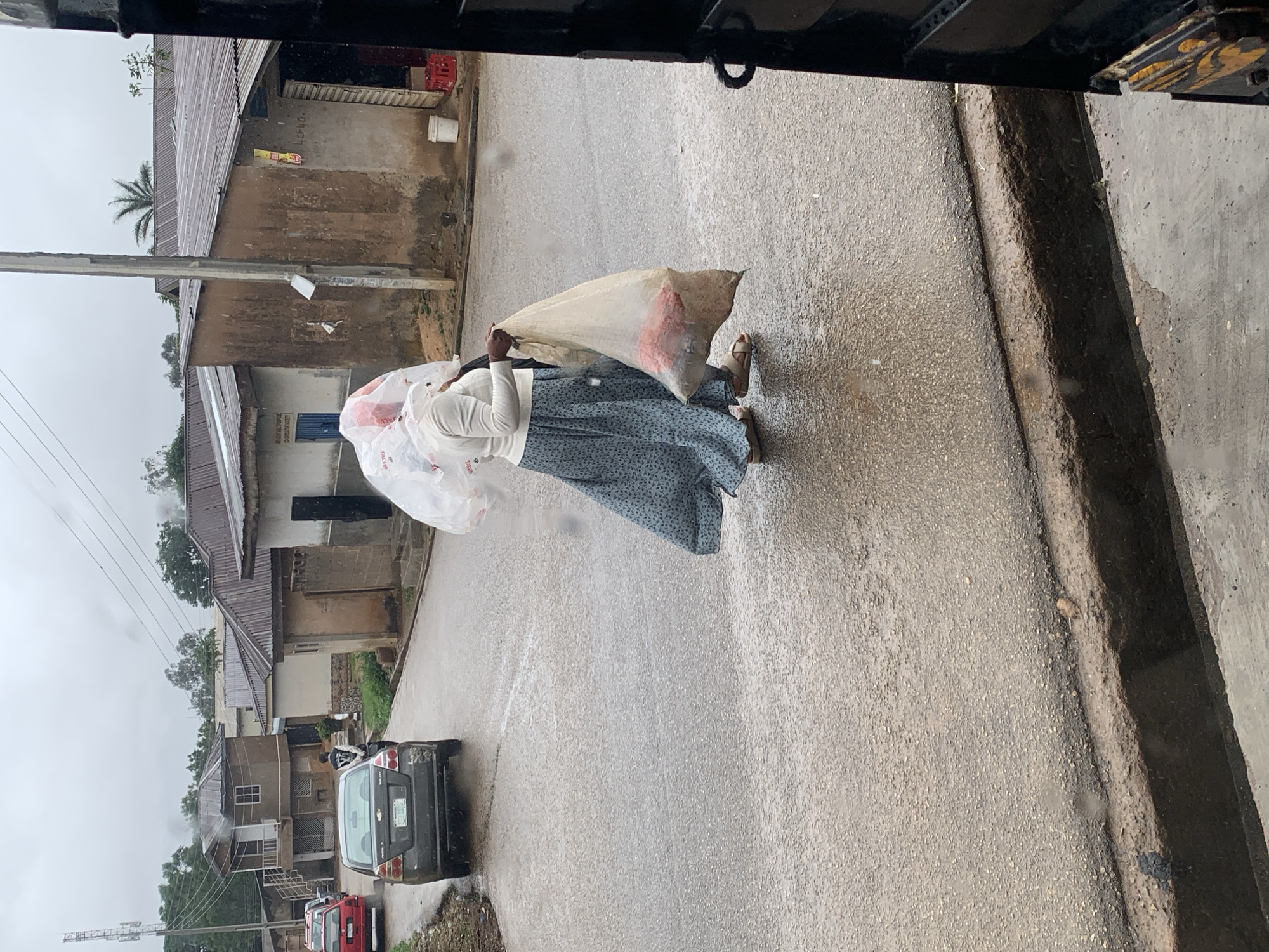 This vertical image captures a street scene in what appears to be a residential or mixed-use area, likely in Jos, Nigeria. The ground is wet, suggesting it has recently rained or is currently raining.
The main subject is a person, likely a woman, walking away from the camera. She is dressed in a white long-sleeved top and a blue-and-white patterned long skirt, wearing sandals. She carries a large, light-colored sack over her right shoulder and holds a translucent white plastic bag in her left hand, which she uses to partially shield her head. The word "NIKE" is partially visible on the plastic bag.
The street is paved, and a concrete ditch or gutter runs along its right edge. Buildings with corrugated metal roofs and concrete or mud-brick walls line the left side of the road. Utility poles and power lines are visible, extending into the distance. Palm trees and other vegetation are scattered throughout the background.
Further down the street, several vehicles are parked, including a dark-colored Chevrolet SUV, a red car, and a white motorcycle. Visible text on the buildings includes "LUDHO" on a yellow sign and "WE ARE NOT WORTHY OF CHRISTIAN GODEY" written above a blue door. The scene depicts a mundane moment of daily life, likely under overcast skies or during daytime after rain.