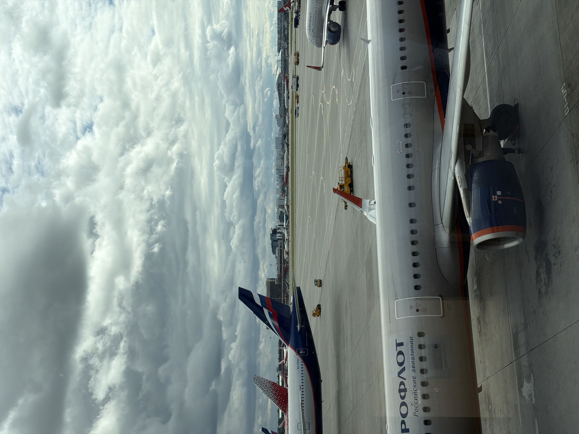This image depicts a view from an airplane window at Sheremetyevo International Airport in Khimki, Russia. The foreground is dominated by the fuselage and wing of a silver Aeroflot airplane, featuring the airline's logo and "Russian Airlines" in Cyrillic script. The aircraft's engine, with its characteristic blue and orange banding, is visible on the right. Beyond this, the tarmac stretches out, populated by several other aircraft of various airlines, including one with a red and white patterned tail. In the mid-ground, ground support vehicles, including yellow baggage tractors, are present. The background reveals the airport terminal buildings and more distant aircraft. The sky is overcast with dramatic, dark clouds. The overall scene is typical of an active airport tarmac under a cloudy sky.