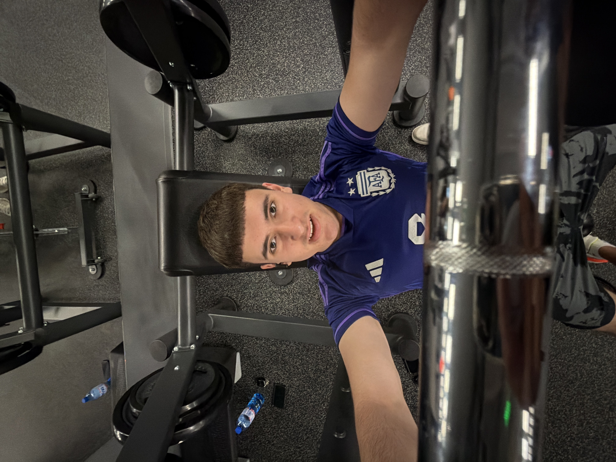 A young man is lying on a weight bench in a gym, likely within the city of Plovdiv, Bulgaria, though direct evidence is lacking. He is wearing a blue soccer jersey with a white insignia on the left side, and he is facing the camera with a slight smile. He is holding a weightlifting bar with his arms extended, likely about to perform a bench press. The gym setting is apparent, with weight plates visible on the bench press machine. There are also water bottles on the ground near the equipment. The floor has a speckled, dark gray texture.