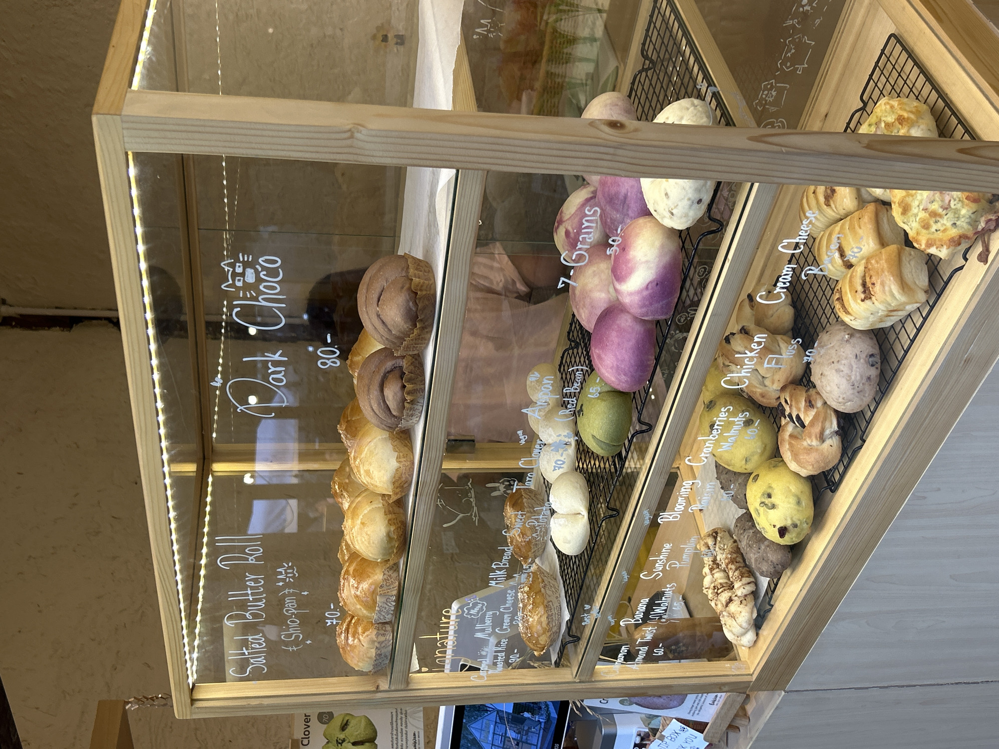 This image shows a close-up view of a bakery display case filled with various baked goods. The display case is made of light-colored wood and has glass fronts. It is illuminated by strings of warm white LED lights.
The subjects are the baked goods, which include a variety of breads and pastries. Some are round, while others are twisted or have intricate shapes. There are also some round, colored items that appear to be buns or sweet breads, with colors like purple and yellow visible. Written on the glass in white chalk are names and prices of the items, such as "Salted Butter Roll," "Dark Choco," "7-Grains," "Cream Cheese," and "Chicken." Prices are also listed, with numbers like "70," "80," and "65" visible.
The scene is a retail setting, likely a bakery or a cafe, given the display of food and the chalk labeling. The background is a textured, light-colored surface, possibly a wall or a countertop. The lighting suggests it might be indoors, perhaps during the day or in the evening when artificial light is used.
There are no people directly visible in the image, but the presence of a "TIP BOX" in the lower right corner suggests customer interaction. The overall impression is one of a well-organized and inviting food display. The location context of Ban Sai Kham, Thailand, doesn't provide specific visual cues within the image itself but sets the geographical context for the establishment.