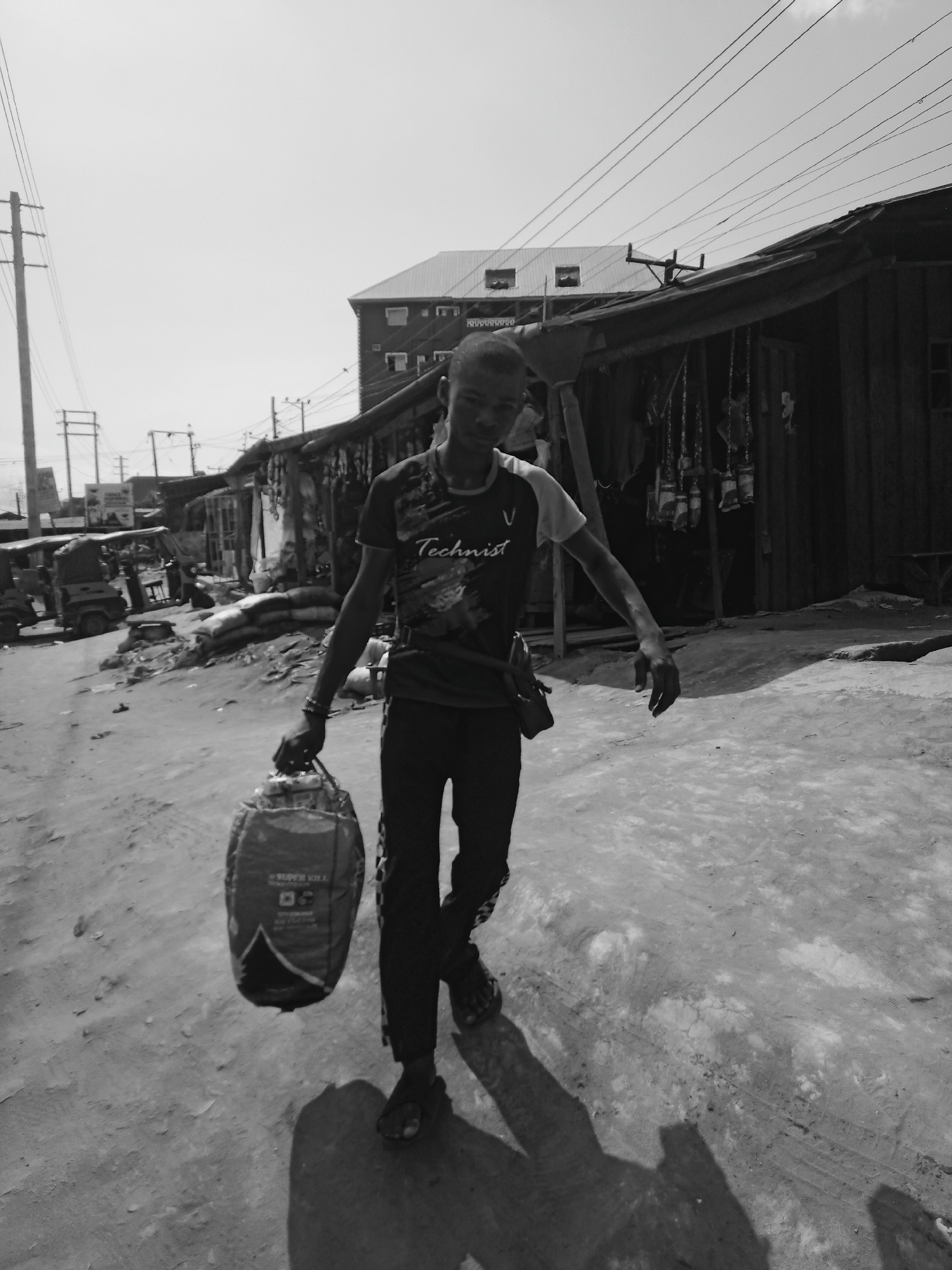 A black and white photograph captures a young man walking along a dusty, unpaved street in Onitsha, Nigeria, under a bright, clear sky. The man, positioned centrally and walking towards the viewer, wears a dark t-shirt with the word "Technist" and a 'V' logo, along with dark trousers. He carries a large, cylindrical bag in his right hand, which appears to be a product sack with the visible text "SUPER KILL" and other symbols.
Strong sunlight from a high angle casts distinct, elongated shadows of the man and nearby objects onto the ground, indicating midday or early afternoon. The street is lined with simple, possibly makeshift, structures and shops, some with corrugated iron roofs and goods hanging outside. Utility poles and a dense network of power lines crisscross the upper part of the image, suggesting an urban or semi-urban setting.
Further down the street, other vehicles, possibly tricycles, and piles of sacks or debris are visible. The overall scene depicts a busy, everyday environment, characteristic of a commercial or residential area in Onitsha, with the man engaged in a routine activity, likely carrying goods or running an errand. The unpaved ground and informal structures convey a sense of bustling daily life.