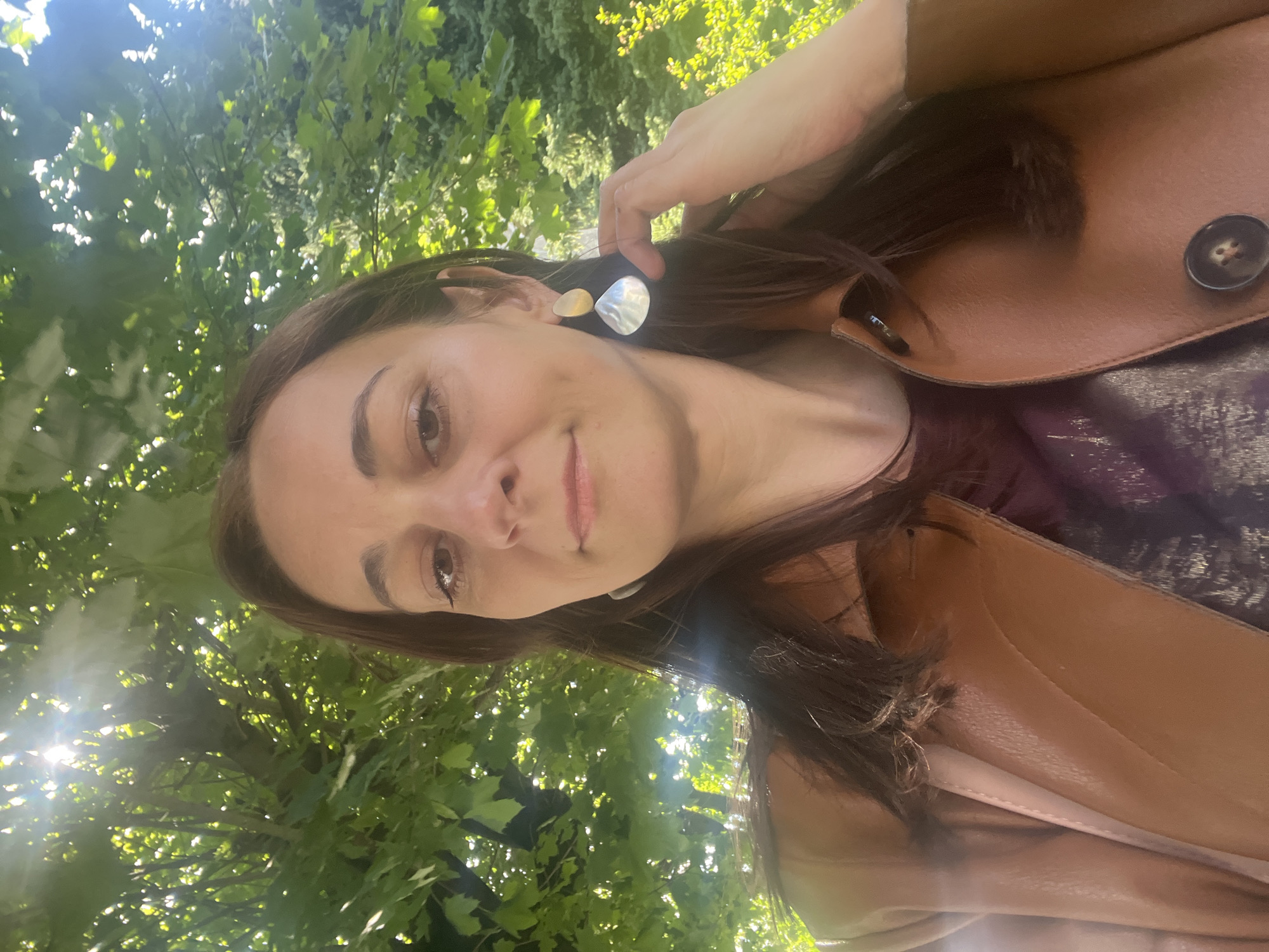 In an outdoor setting bathed in sunlight, a woman with long, dark brown hair takes a selfie. She is wearing a brown leather jacket over a shimmering dark purple shirt. Her makeup is subtle, with mascara and eyeliner defining her eyes. She has on large, unique earrings; one is a golden-toned teardrop shape, and the other is a larger, iridescent silver shell-like shape. Her right hand is raised to her hair, and her fingers are gently touching her earlobe, close to the earring. The background is filled with lush green foliage, suggesting trees and shrubs, with bright sunlight filtering through the leaves. The scene appears to be in a park or garden.